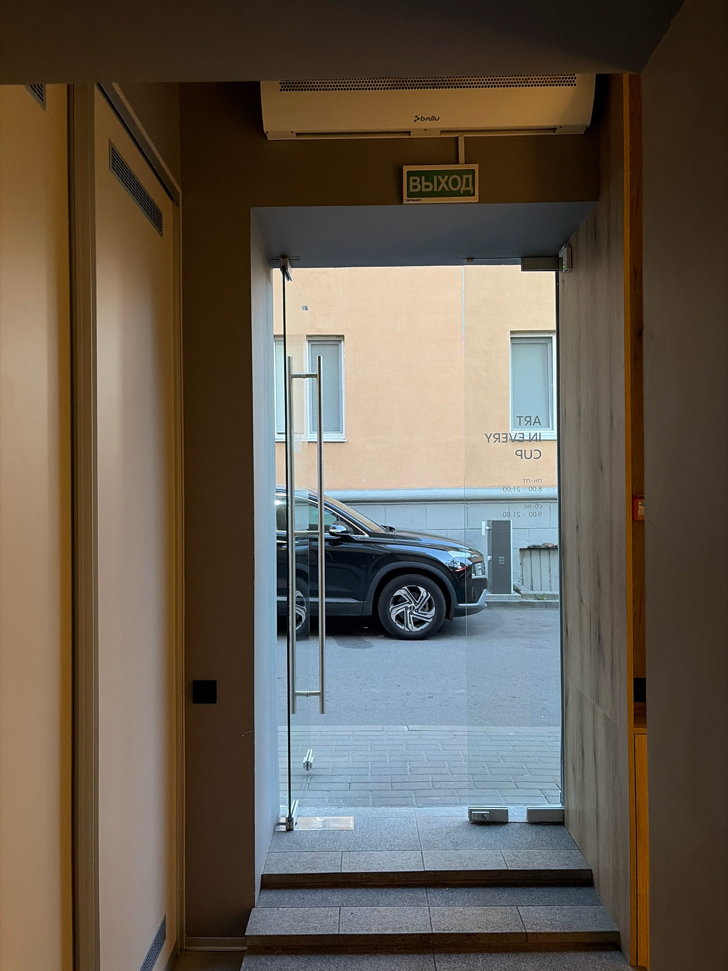 The image shows an entrance to a building with a glass door. Above the door, there is an air conditioner and an exit sign written in Russian, "ВЫХОД" (VYKHOD), which translates to "EXIT". Through the glass door, a black SUV is visible parked on the street. On the glass door, there is lettering that appears to be reversed, reading "ART IN EVERY CUP". Below this, there are also reversed numbers that seem to represent operating hours, such as "00.8 - 51.00" and "00.8 - 51.00". The building across the street has a light-colored facade and windows. The overall setting suggests an urban environment. Given the Russian text on the exit sign and the lettering on the glass, the location is likely in a Russian-speaking country. While the prompt specifically asks for Minsk, Belarus, there are no definitive visual cues in the image to confirm this specific location beyond the implied Eastern European context due to the Russian language.