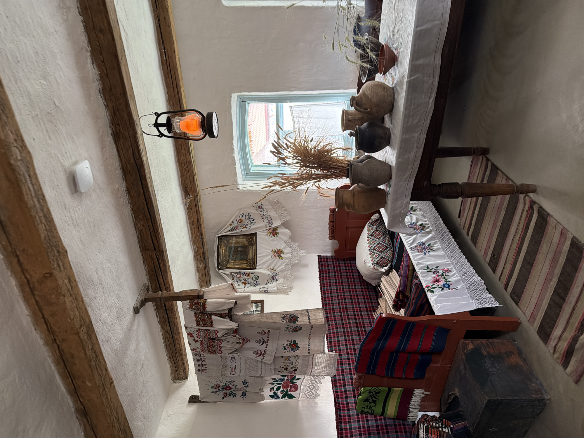 This image depicts a rustic interior, likely within a dwelling in Velykyi Khodachkiv, Ukraine. The room features exposed wooden beams on the ceiling and whitewashed walls. A red-framed bed is partially visible, adorned with plaid fabric and embroidered linens, including a pillow with geometric patterns. To the left of the bed is a dark wooden chest.
A long wooden table is covered with a white cloth, upon which several clay pots and vases are arranged, alongside a bouquet of dried wheat stalks. Further down the table, more ceramic vessels and greenery are present. A small, reddish-brown bowl sits on the table to the right.
Hanging on the wall above the bed are several pieces of embroidered fabric, likely towels or decorative cloths, with intricate floral and geometric designs. A framed image is also hung on the wall to the right of these textiles.
A kerosene lantern with an illuminated orange glow hangs from the ceiling in the center of the room. A window with a light blue frame is visible in the background, offering a glimpse of an outdoor scene with red-tiled roofs. A striped rug lies on the floor in front of the bed and extends towards the table. The overall impression is one of traditional folk decor and a preserved historical ambiance. There are no people visible in the image.