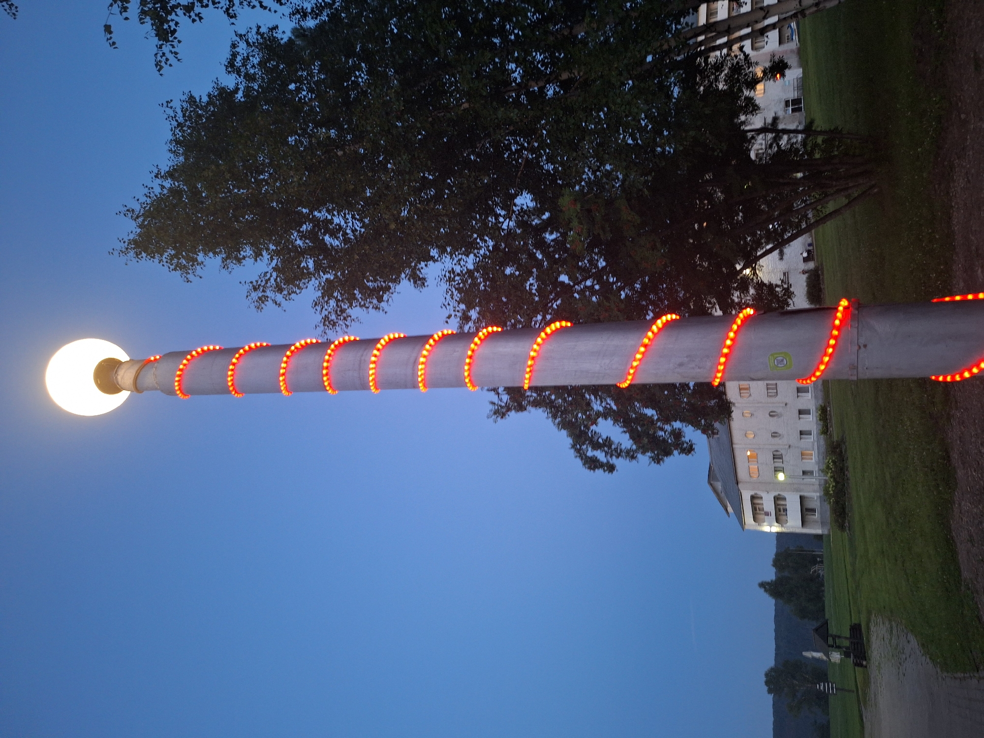The image shows a tall, metal lamppost decorated with a spiral of red LED lights, extending from its base to near the top. A bright white light emanates from the spherical lamp at the very top of the post. The lamppost stands in a grassy area in front of a multi-story building. The building is light-colored with multiple windows, some of which are illuminated. Trees are visible behind and to the right of the lamppost, and a darker treeline is in the background, suggesting a rural or semi-rural setting. A paved pathway leads to the left of the lamppost. The sky is a deep blue, indicating dusk or early evening. The location is identified as Atamanovka, Russia. There are no people or distinct actions depicted in the immediate vicinity of the lamppost.