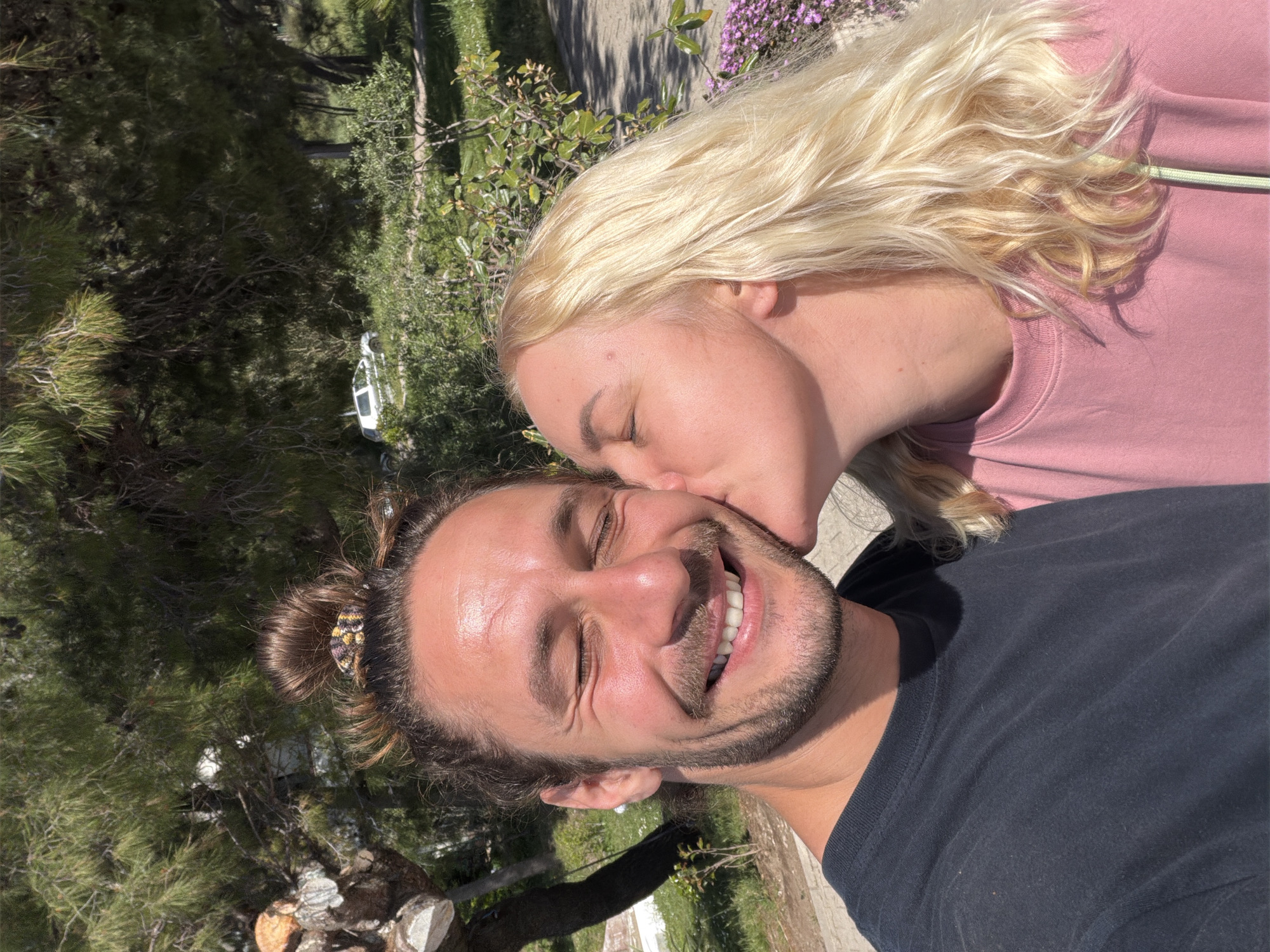 This brightly lit outdoor photograph captures a warm, affectionate moment between a man and a woman in Bar, Montenegro. The man is prominently featured in the foreground, looking towards the camera with his eyes closed and a broad, genuine smile that reveals his teeth. He has dark hair, styled in a bun held by a patterned scrunchie, and a well-groomed beard and mustache. He is wearing a simple black t-shirt.
Behind him, a woman with long, wavy blonde hair leans in to kiss his right cheek. Her eyes are also closed, suggesting a moment of tenderness and contentment. She is dressed in a light pink long-sleeved top, with a light green drawstring visible near her neckline.
The background is characterized by abundant natural greenery, including tall pine trees and various bushes, indicating a sunny, pleasant day. Some small patches of purple flowers add a touch of color to the verdant setting. A paved path or road is visible, and a white vehicle can be faintly seen among the trees in the middle distance. The clear, strong sunlight casts defined shadows, typical of mid-day or early afternoon. The overall mood conveyed is one of happiness and connection. No text is visible in the image.