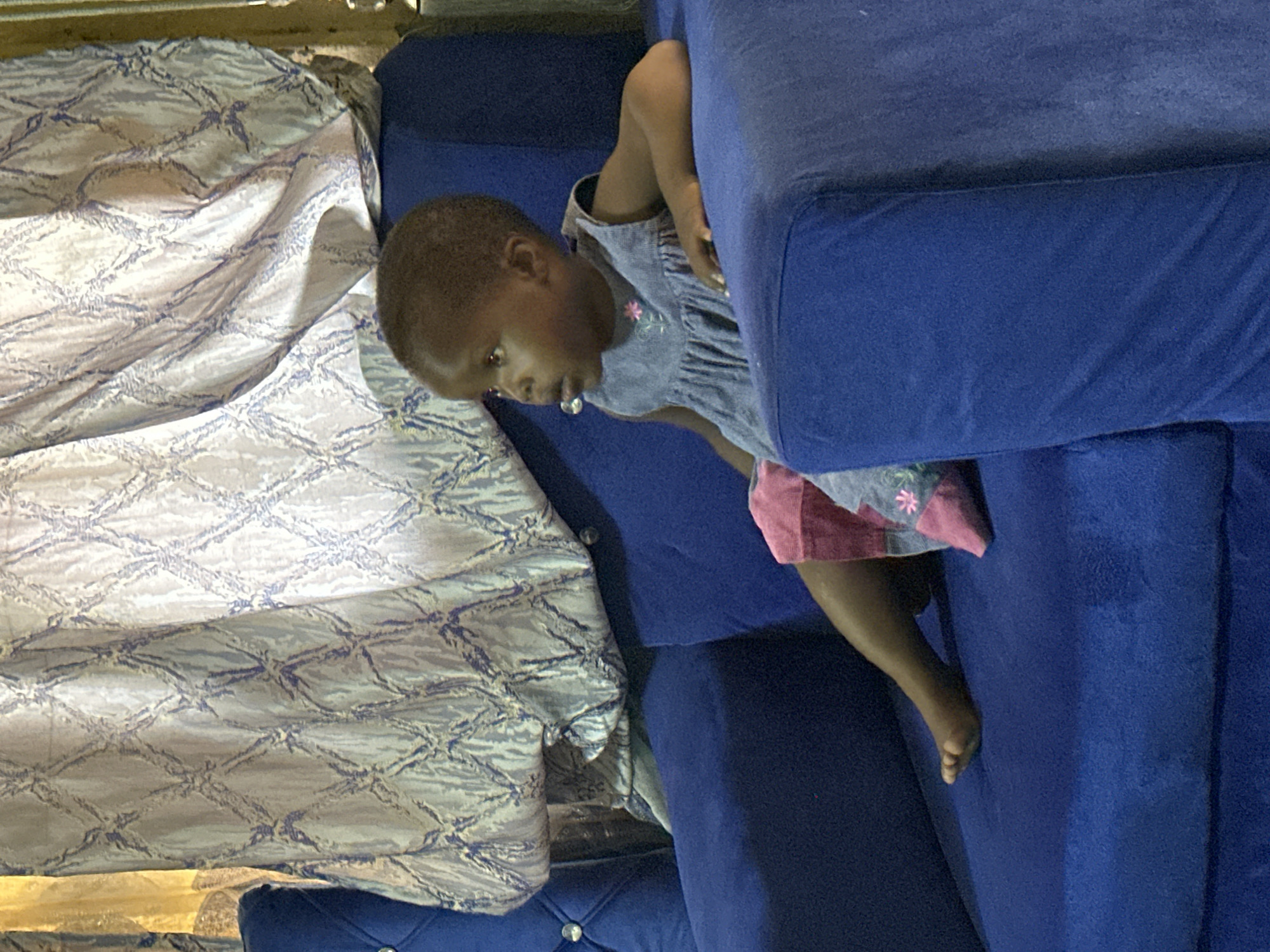 A young child is seated on a blue couch in Narayi, Nigeria. The child, wearing a light blue sleeveless dress with a pink floral appliqué, is looking to the right. The couch is upholstered in a textured blue fabric. Behind the child, a patterned curtain with a diamond motif in blue and off-white hangs.
