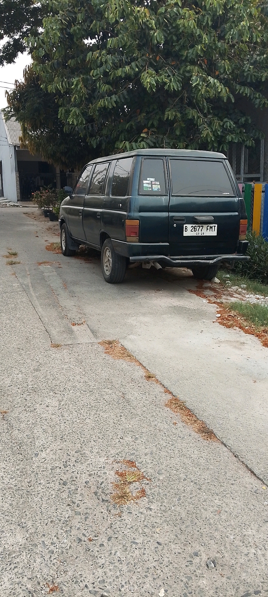 An external, eye-level, full shot captures a dark green Isuzu Panther van parked on an asphalt road with patches of dried grass and debris. The van, facing right, is viewed from its rear-left quarter. The license plate reads "B 2677 FMT" with smaller text below. The van has tinted windows, some with stickers. To the left of the van, a gray and white house with a dark roof is visible, bordered by potted plants. Behind the house, a large tree with green leaves dominates the background, with its branches extending over the road and van. The environment appears to be a residential area in Satriamekar, Indonesia, indicated by the architectural style of the house and the vehicle's license plate.