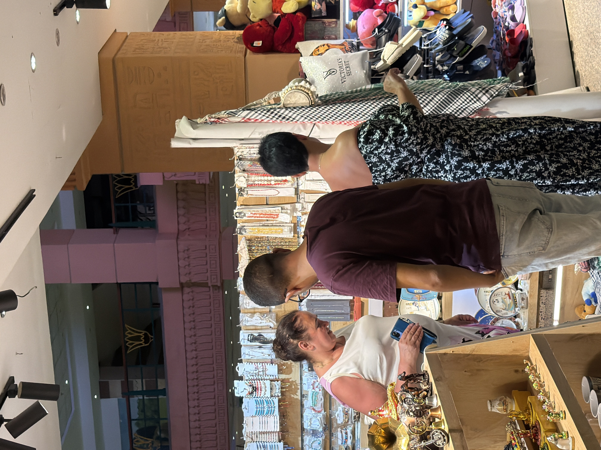 In Sharm El Sheikh, Egypt, three individuals are observed inside a marketplace or souvenir shop. The woman on the left, with dark hair pulled back, wears a white top and is looking down at her phone, which she holds in her right hand. Next to her, a man with short hair and glasses, dressed in a maroon t-shirt and light-colored jeans, faces away from the camera and appears to be looking at merchandise. A second woman, with short dark hair, stands to the right of the man, wearing a black and white floral off-the-shoulder dress. She is reaching out to examine items on display. The background reveals shelves stocked with various souvenirs, including jewelry, decorative plates, and plush toys, with Egyptian-themed architectural elements visible on pillars. The overall setting suggests a retail environment focused on tourist goods.