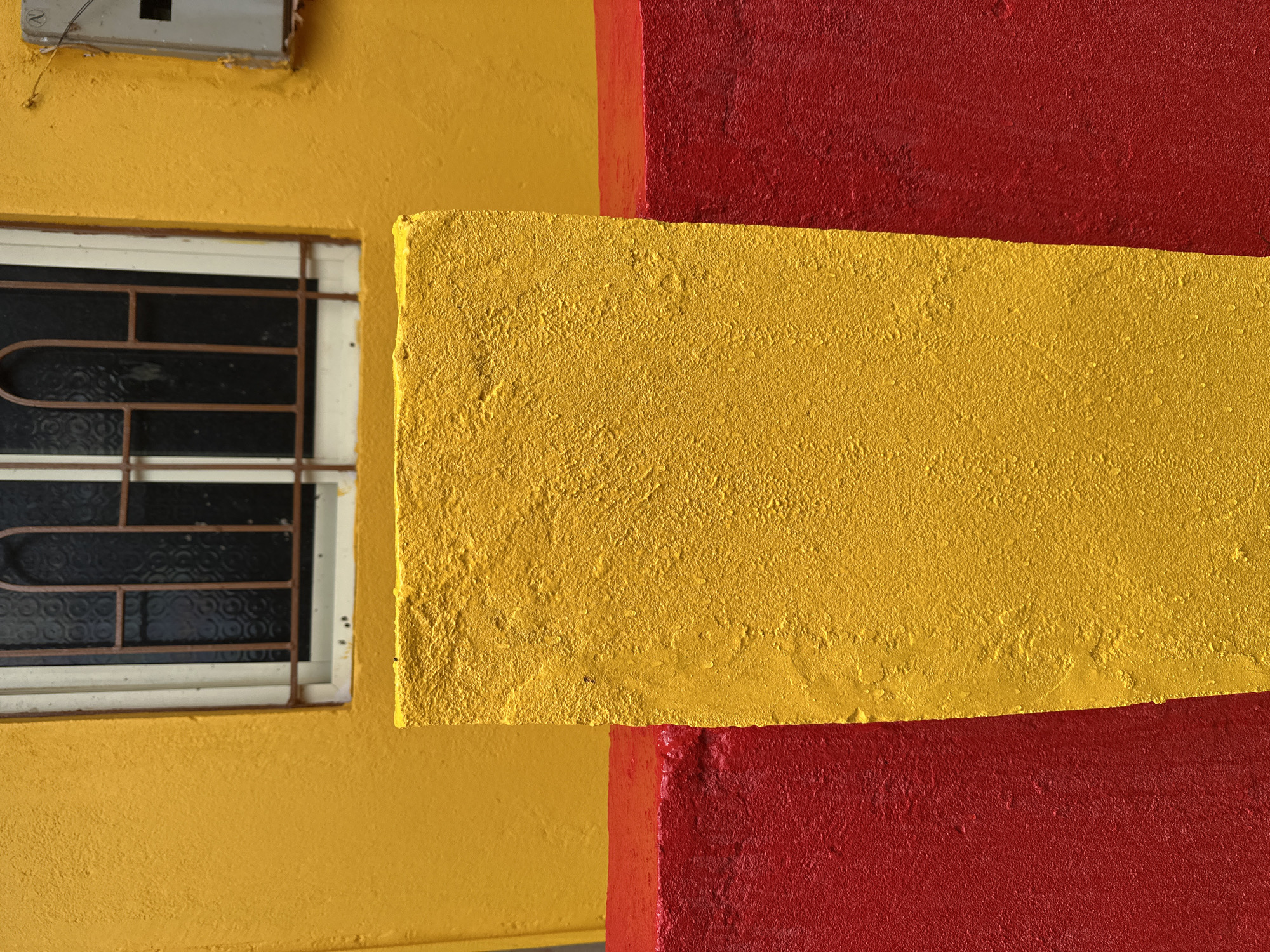A close-up, eye-level shot captures a portion of a building's exterior in Arambol, India. The dominant colors are bright yellow and red, creating a bold visual contrast.
In the foreground, a textured, yellow rectangular pillar stands prominently, centered in the frame. This pillar is situated on top of a horizontal, textured red surface that extends across the bottom half of the image. The red surface appears to be a raised border or ledge.
Behind the yellow pillar, another horizontal red surface is visible, creating a layered effect. The building wall behind these elements is painted a vibrant yellow. A small, barred window is set into the yellow wall. The window has a dark, patterned glass and a simple metal grate. To the right of the window, a small, light-colored electrical box or meter is attached to the wall. The overall scene is stark and abstract, focusing on color and texture. There are no people or visible actions taking place. The environment appears to be outdoors, with the bright colors suggesting a sunny day.