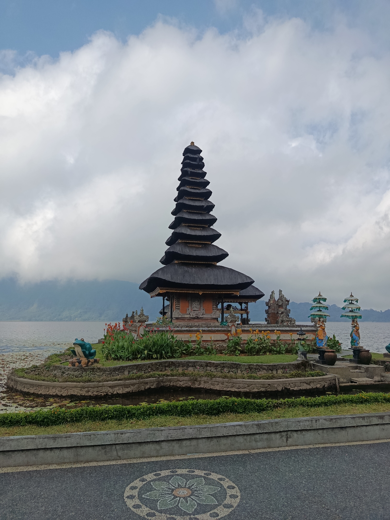 A Balinese temple complex, Pura Ulun Danu Bratan, is situated on the edge of Lake Batur. The temple features a multi-tiered pagoda-style roof and is surrounded by lush greenery and decorative statues. The lake is calm and reflects the cloudy sky. In the foreground, a paved pathway with a mosaic design leads towards the temple. The scene is tranquil and serene, with a hint of mist over the distant mountains. The location is the city of Baturiti, Indonesia.