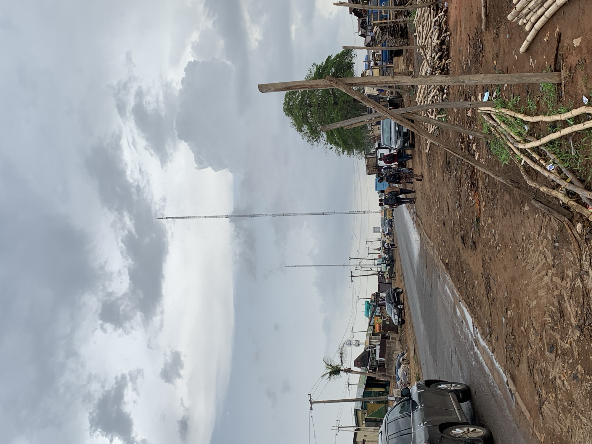 This image captures a wide-angle view of a busy roadside scene in Ikorodu, Nigeria, under an overcast sky with heavy grey clouds. The photograph is oriented vertically, showing a road stretching into the distance with activity on both sides.
In the foreground, a dark SUV is partially visible on the left, parked alongside the paved road. The road itself appears wet or damp in places, and its shoulders are unpaved, consisting of muddy, reddish-brown dirt and debris.
To the right, there are numerous stacks of cut bamboo poles or logs, indicating a potential market or storage area for building materials. Several rough-hewn wooden posts are erected along this side, possibly for temporary structures or stalls. Green foliage and patches of grass grow amidst the dirt and logs.
Along the road, several individuals and small groups of people are walking, suggesting daily pedestrian traffic. Vehicles, including cars and possibly trucks, are visible parked or moving further down the road. Simple, low-rise buildings and structures, likely commercial stalls or residences, line both sides of the street.
Prominently, a very tall, slender communication or radio tower stands in the middle ground, extending high into the cloudy sky. Another, smaller tower is visible further down the road. Power poles and lines run alongside the street. A large, leafy green tree stands out in the middle distance on the right, providing a natural contrast to the built environment.
The scene depicts a typical bustling street in a semi-urban or urban area, characterized by everyday activity, basic infrastructure, and the informal economy suggested by the material piles. No discernible text is clearly visible in the image.