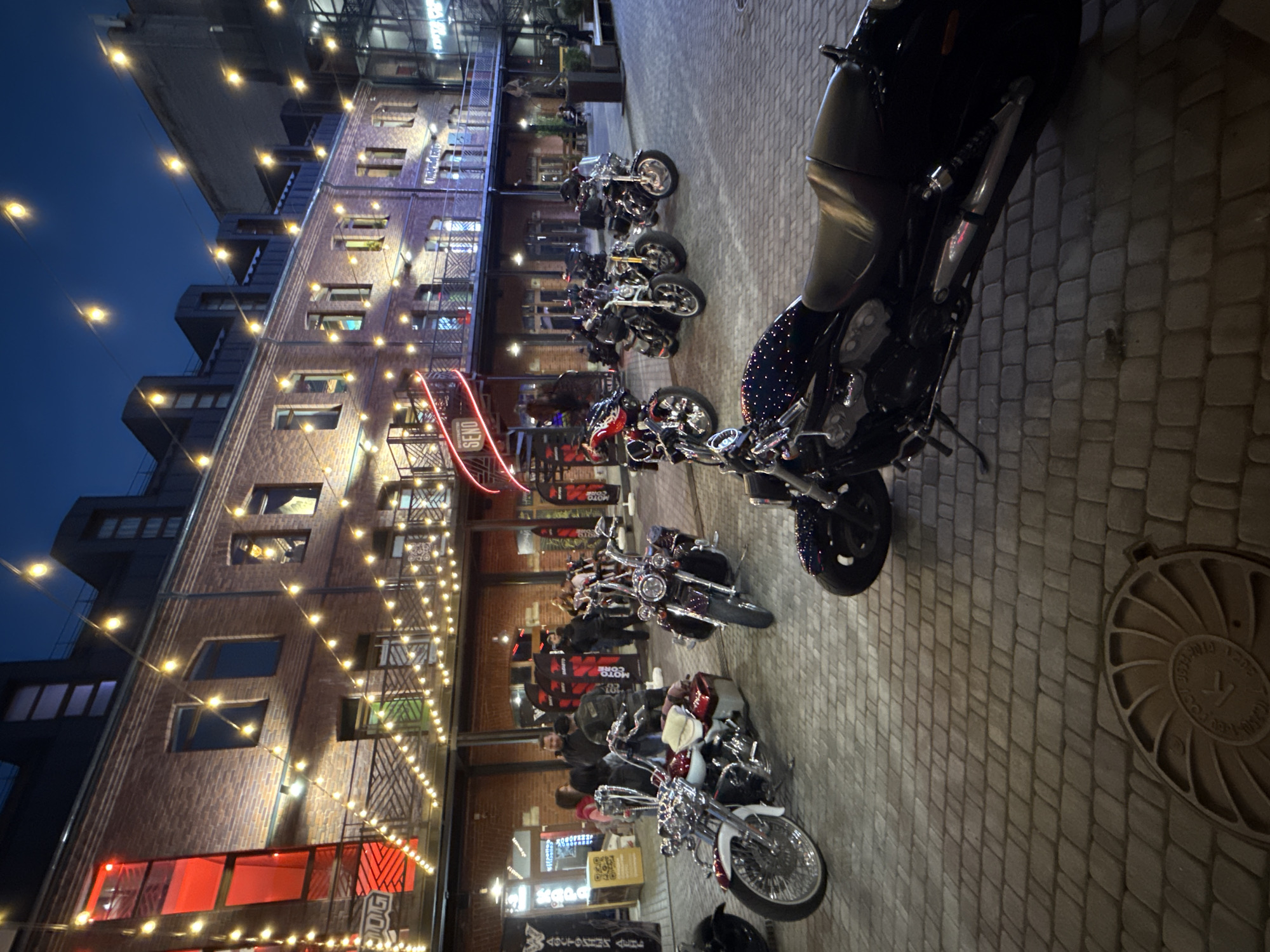 An outdoor night scene in Saint Petersburg, Russia, features a courtyard with several parked motorcycles and a few people milling about. The courtyard is paved with cobblestones. A brick building with multiple stories serves as the backdrop, adorned with strings of warm, glowing fairy lights. Some of the motorcycles are customized, with chrome accents and intricate designs. The overall atmosphere is one of a casual gathering or event, possibly related to motorcycle culture. The building's architecture suggests an older, possibly repurposed industrial or historical structure. The sky above is dark, indicative of nighttime.