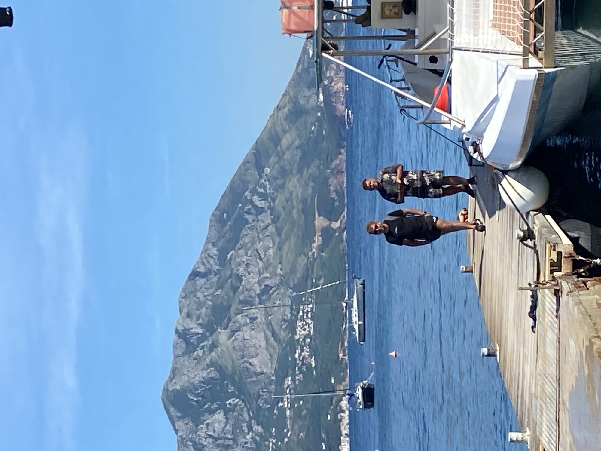 Two men are standing on a wooden pier in Budva, Montenegro. The man on the left is wearing a black t-shirt and black shorts, and the man on the right is wearing a patterned short-sleeved shirt and camouflage shorts. They are both looking towards the camera. In the background, there are several sailboats anchored in the blue water. A large, green, mountainous island with buildings on its slopes is visible behind the sailboats. The sky is clear and blue. A white boat with a red life preserver is docked on the right side of the frame.