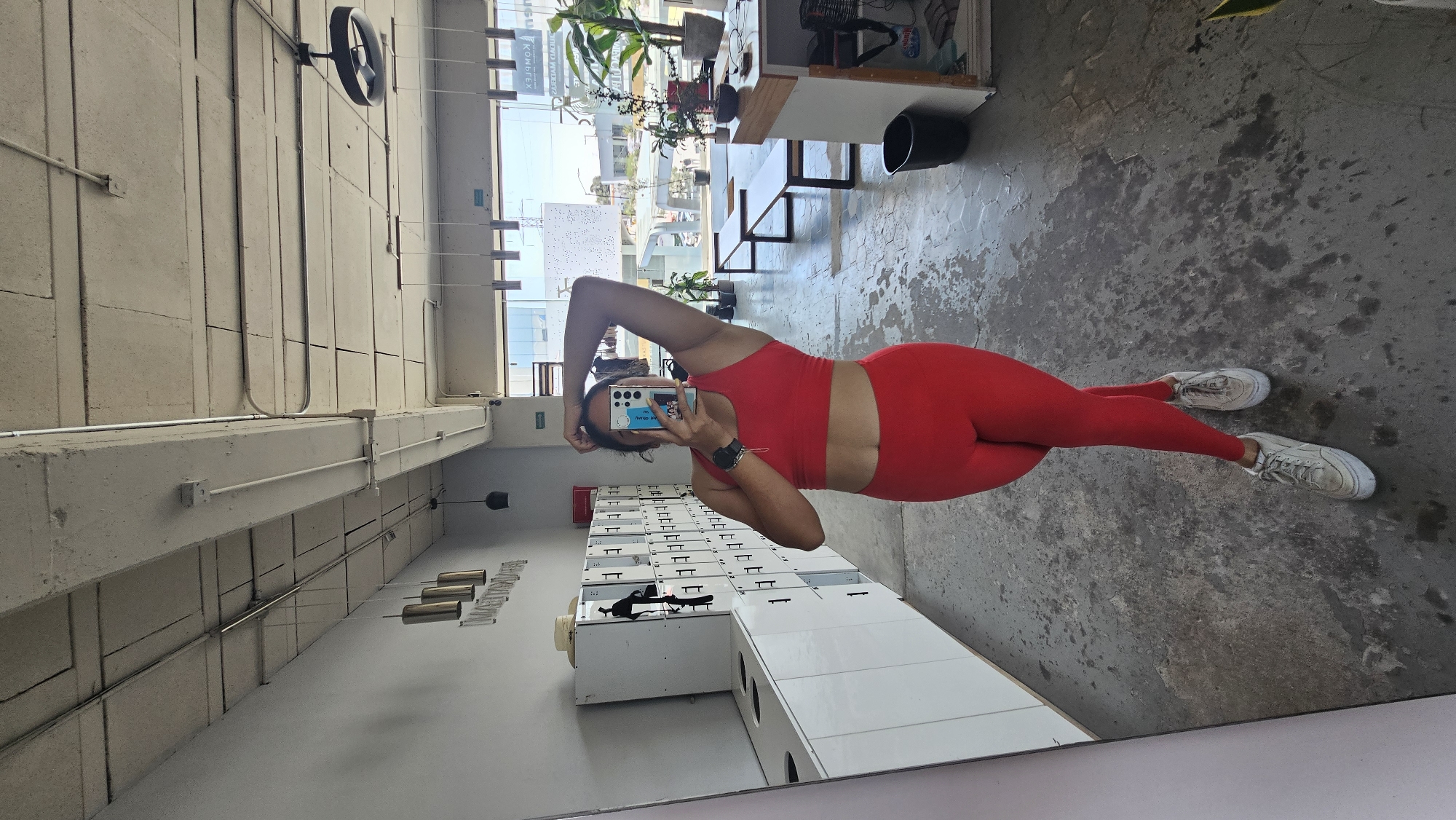 A woman in red athletic wear stands in front of a mirror, taking a selfie. The background shows a gym setting with lockers, exposed ceiling beams, a fan, and a window showing a street scene in Puebla, Mexico. In the window, blurred buildings and greenery are visible. The floor is concrete.