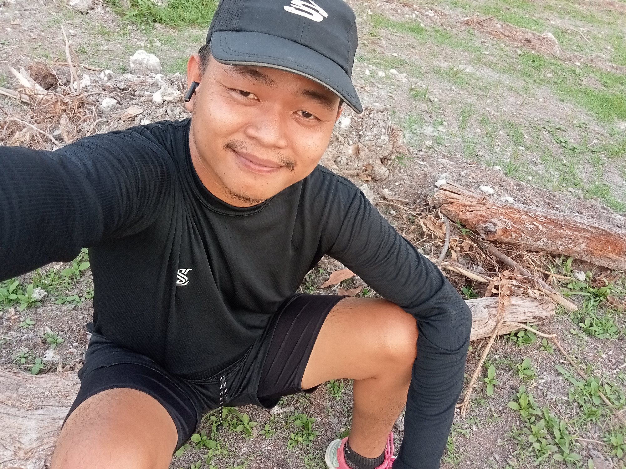 A man is taking a selfie outdoors. He is wearing a black baseball cap, a black long-sleeved shirt, and black shorts. He has a mustache and is smiling at the camera. He is sitting on the ground amidst dry twigs and small plants. In the background, there is more dry foliage and patches of grass. A piece of weathered wood is visible in the foreground. The man is wearing an earbud in his left ear and a pink and white athletic shoe on his right foot. The location is the city of Teluk Kenidai, Indonesia.