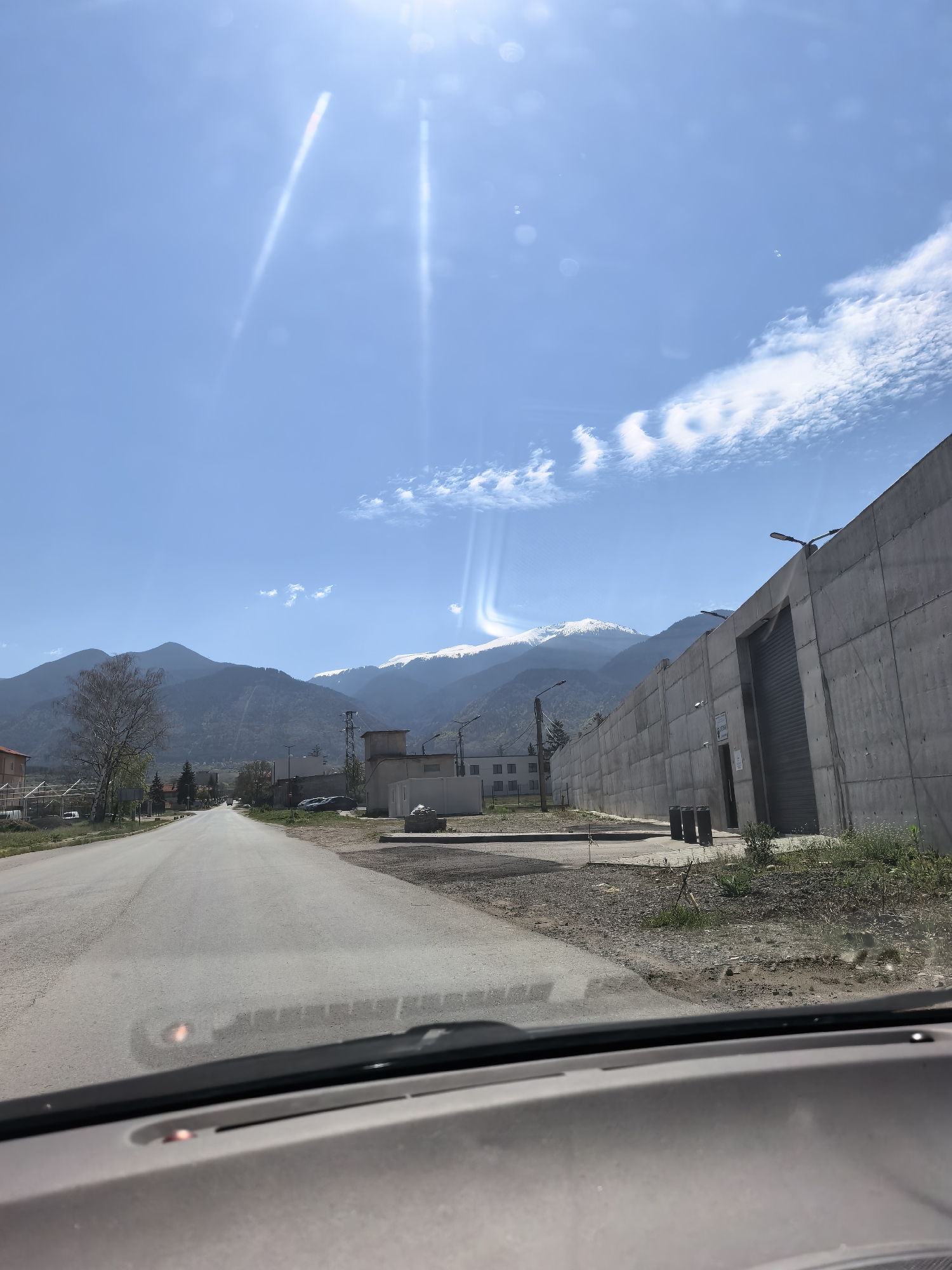 The image captures a sunny daytime view from inside a car, looking down a straight road towards distant snow-capped mountains. The perspective is through the front windshield, with the dashboard visible in the foreground.
On the left side of the road, there's a small village or town with scattered buildings, including a prominent bare tree with some green foliage. Utility poles and lines run alongside the road. The immediate roadside on the left has some sparse grass and dirt.
To the right of the road, a large, tall concrete wall dominates the midground, suggesting an industrial or secure facility. This wall features a large, dark metal rolling door and several streetlights mounted along its top edge. Near the wall, there are three dark grey bins and some overgrown weeds and dirt. A small white sign is affixed to the concrete wall, but the text on it is illegible.
In the background, a range of mountains rises, with the peaks further back covered in snow. Closer mountains are dark green/grey, indicating forested or rocky terrain. The sky is clear blue with some wispy white clouds, particularly towards the right. Bright sunlight causes significant lens flares and sunbeams, especially in the upper left and center-right of the sky, indicating the sun's position.
No people are visible in the scene. The activity depicted is traveling or being stationary in a vehicle on a clear, bright day. The overall impression is one of a journey through a rural or semi-urban area with striking natural scenery in the distance.