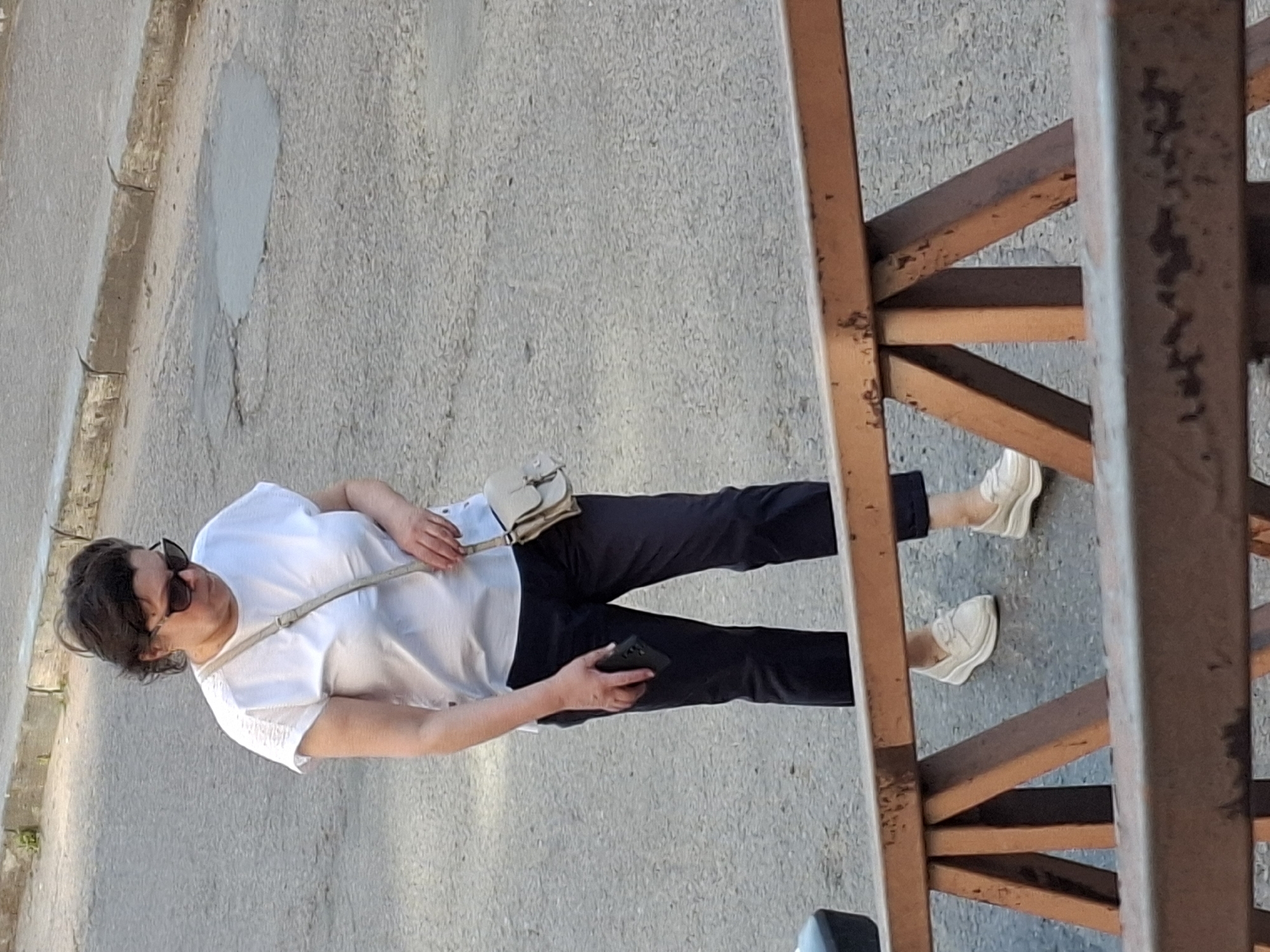 A woman with dark, curly hair and sunglasses stands outdoors, looking down. She is wearing a white t-shirt, dark pants, and light-colored slip-on shoes. A beige crossbody bag is slung over her right shoulder. She is holding a dark-colored smartphone in her left hand. The ground is paved with asphalt, with some cracks and patches visible. A rusty metal barrier is in the foreground. The scene appears to be on a street.