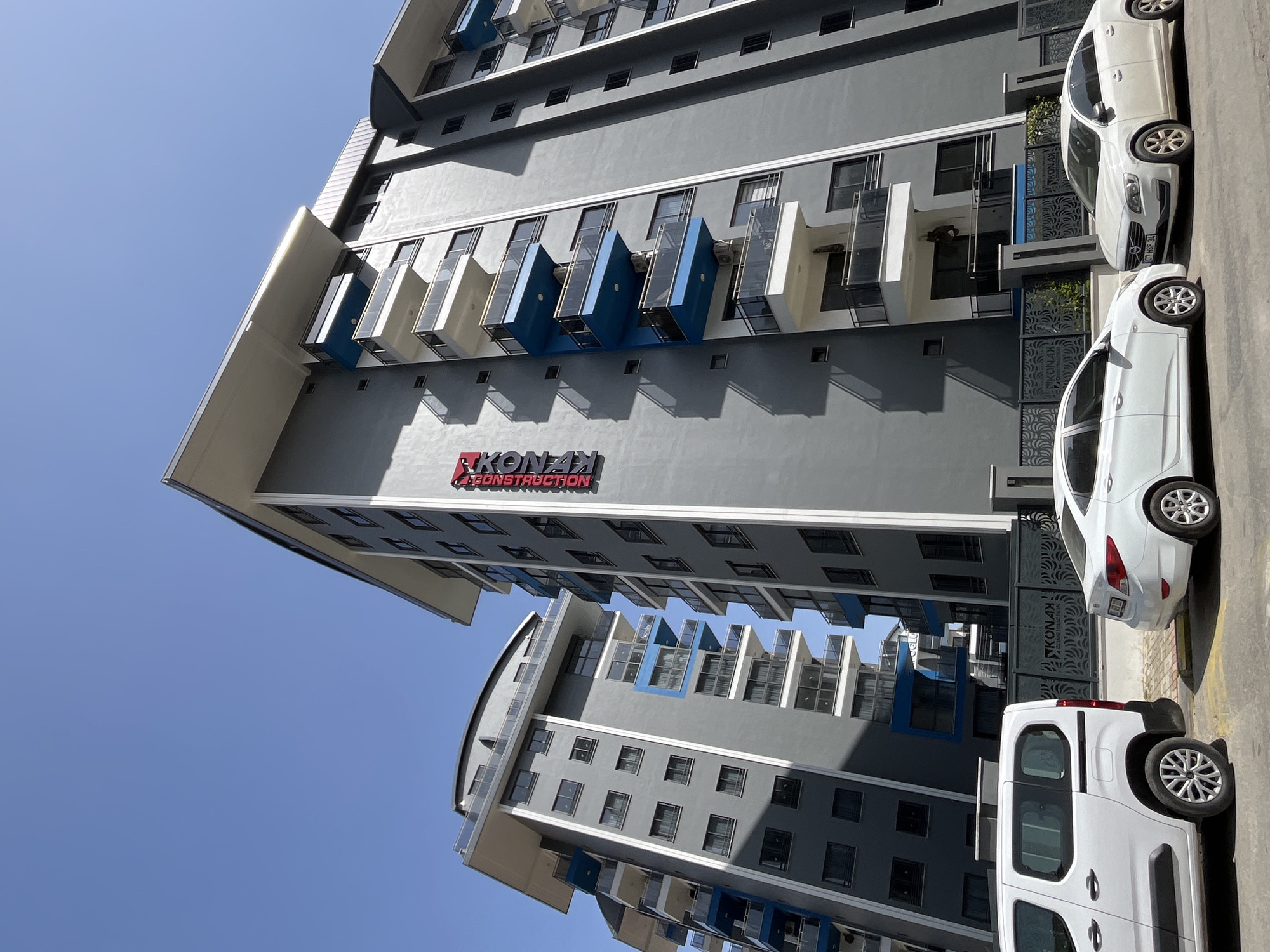 The image shows a multistory gray building under a clear blue sky, located in Alanya, Turkey. The building features projecting balconies painted white and blue, with multiple windows on each floor. A large sign reading "RKONAK CONSTRUCTION" in red is visible on the facade of the building. Three white vehicles are parked on the right side of the frame, next to a gray fence with a decorative pattern. The architectural style suggests a contemporary design. The photograph is taken from a low angle, emphasizing the height of the building.