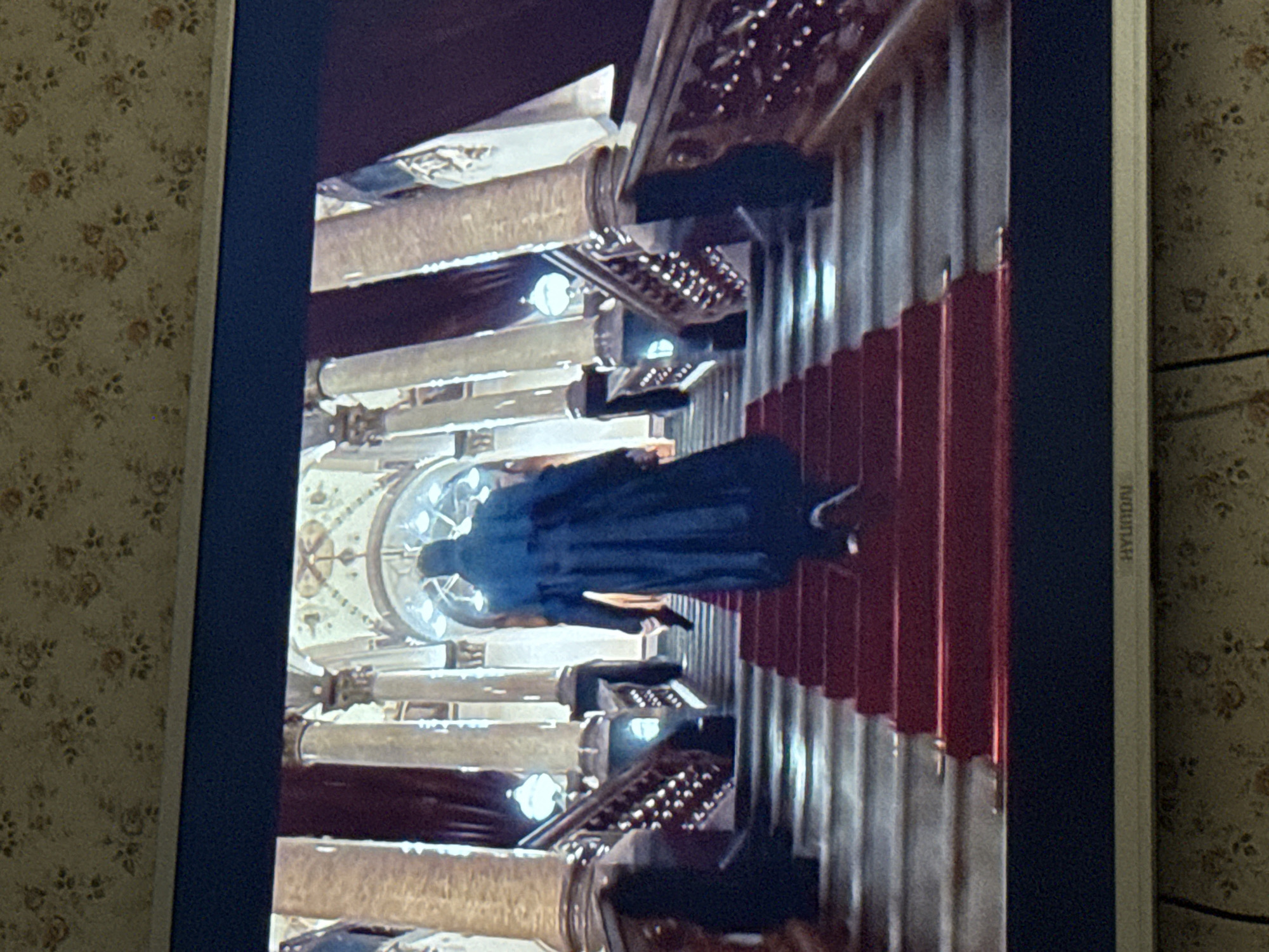 A television displays a scene of a person in dark clothing walking down a red-carpeted staircase in an ornate, palatial interior. The architecture features detailed pillars, arches, and a domed ceiling with an intricate design. The scene evokes a sense of grandeur and formality. No location details are visible in the image.