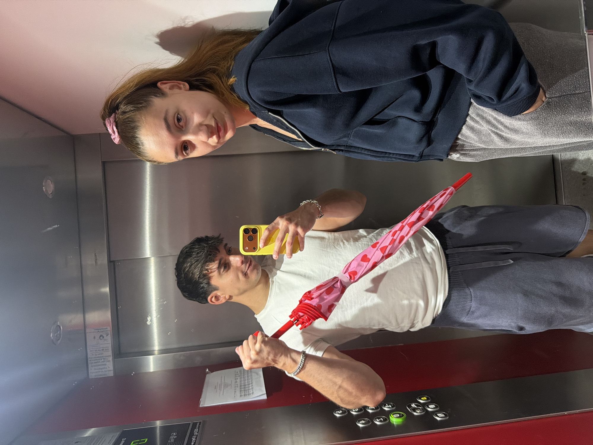 Two individuals are inside an elevator. A man is holding a pink and red heart-patterned umbrella and a yellow smartphone, seemingly taking a photo. The woman is wearing a navy blue hoodie and gray pants. The elevator's interior is primarily silver, with red accents near the floor. Elevator buttons and a control panel are visible. The location is indicated to be Sofia, Bulgaria.