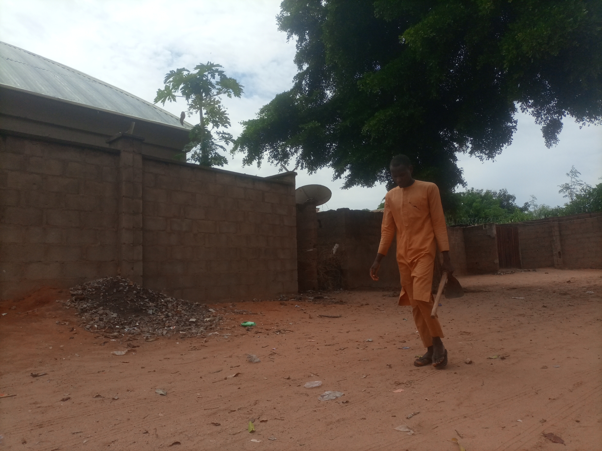 In Tumfure, Nigeria, a lone man dressed in an orange kaftan walks on a dirt path. He carries a long-handled tool, possibly a hoe, in his right hand. The path is a reddish-brown dirt, scattered with debris. To the left, a brick wall stretches across the frame, with a pile of rubble in front of it. A building with a corrugated metal roof is visible behind the wall. Large trees with dense foliage dominate the background, casting shadows on the scene, and the sky is overcast. A satellite dish is mounted on the wall.