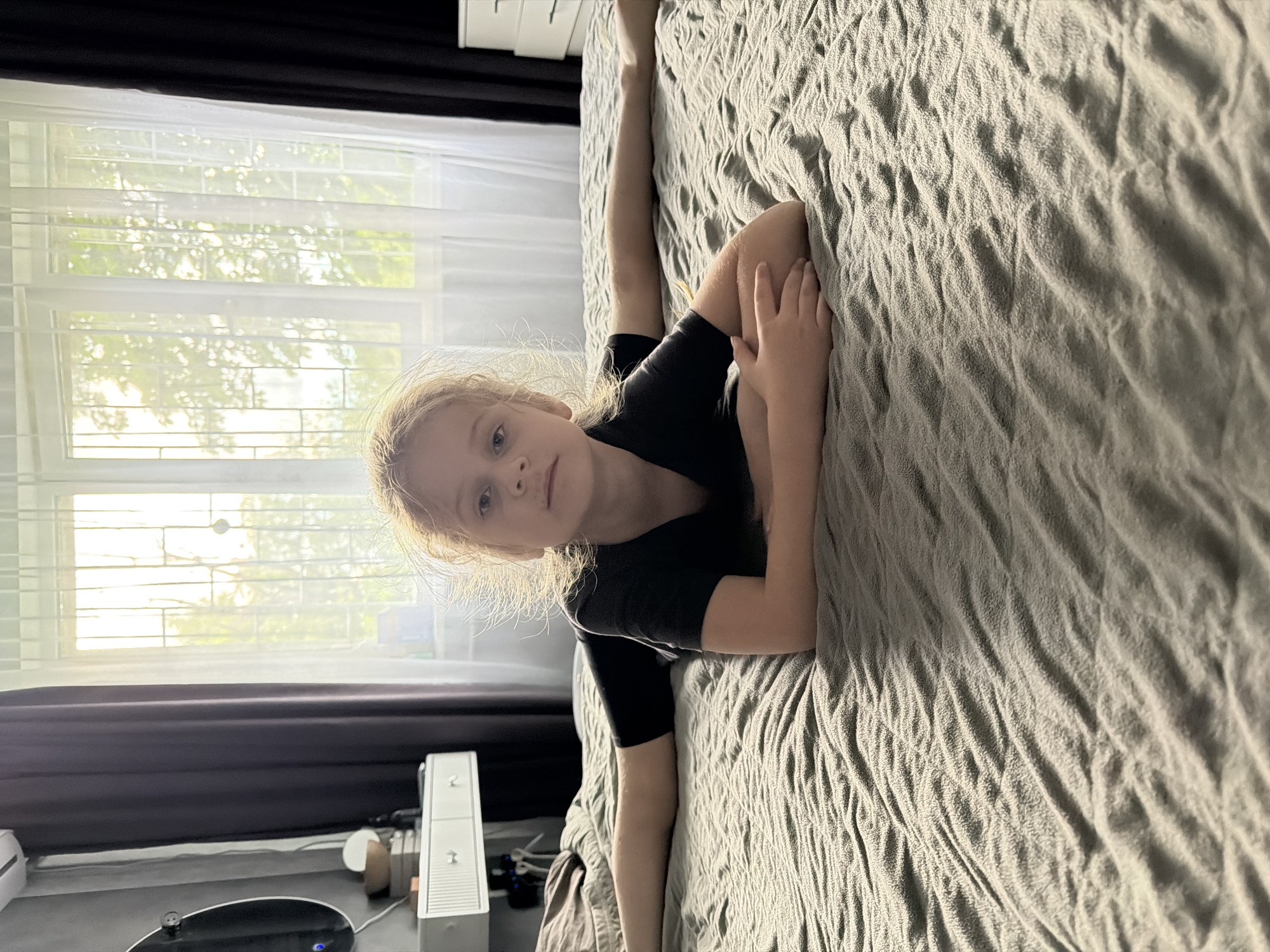 This media file is an image of a young girl with blonde hair lying on a bed. She is wearing a black t-shirt and is positioned in a split, with one leg extended upwards and the other bent. Her face is turned towards the camera, and she has a neutral expression. The bed has a textured, grey comforter. Behind her, a window with blinds is visible, letting in bright, natural light that suggests it is daytime, possibly late morning or early afternoon, given the quality of light. Tree branches are visible through the window, indicating an outdoor setting with foliage. In the lower left portion of the image, a dark surface, possibly a desk or dresser, is visible with some objects on it, including a white electronic device that resembles a gaming console. The overall scene appears to be indoors, in a bedroom or living area.