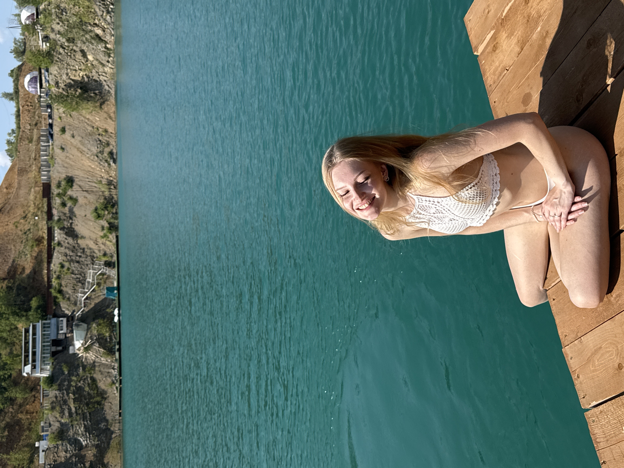 A young woman with blonde hair is seated on the edge of a wooden dock, her legs extended towards the vibrant turquoise water. She is wearing a white crocheted bikini top and bottoms. Her eyes are closed, and she has a joyful smile on her face, suggesting she is enjoying the warm weather and sunny environment.
The scene is set at Karyer Izvestnyak, Russia, where a steep, rocky hillside borders the water. On the hillside, there are several white dome-shaped structures and a modern building with large windows. Stairs are visible leading down the side of the hill towards the water's edge. The water itself appears calm with subtle ripples, reflecting the clear, bright sky. The time of day is daytime, indicated by the strong sunlight casting shadows on the wooden dock. The overall mood is relaxed and pleasant, with the woman appearing to be in a state of happiness and contentment. There is no visible text in the image.