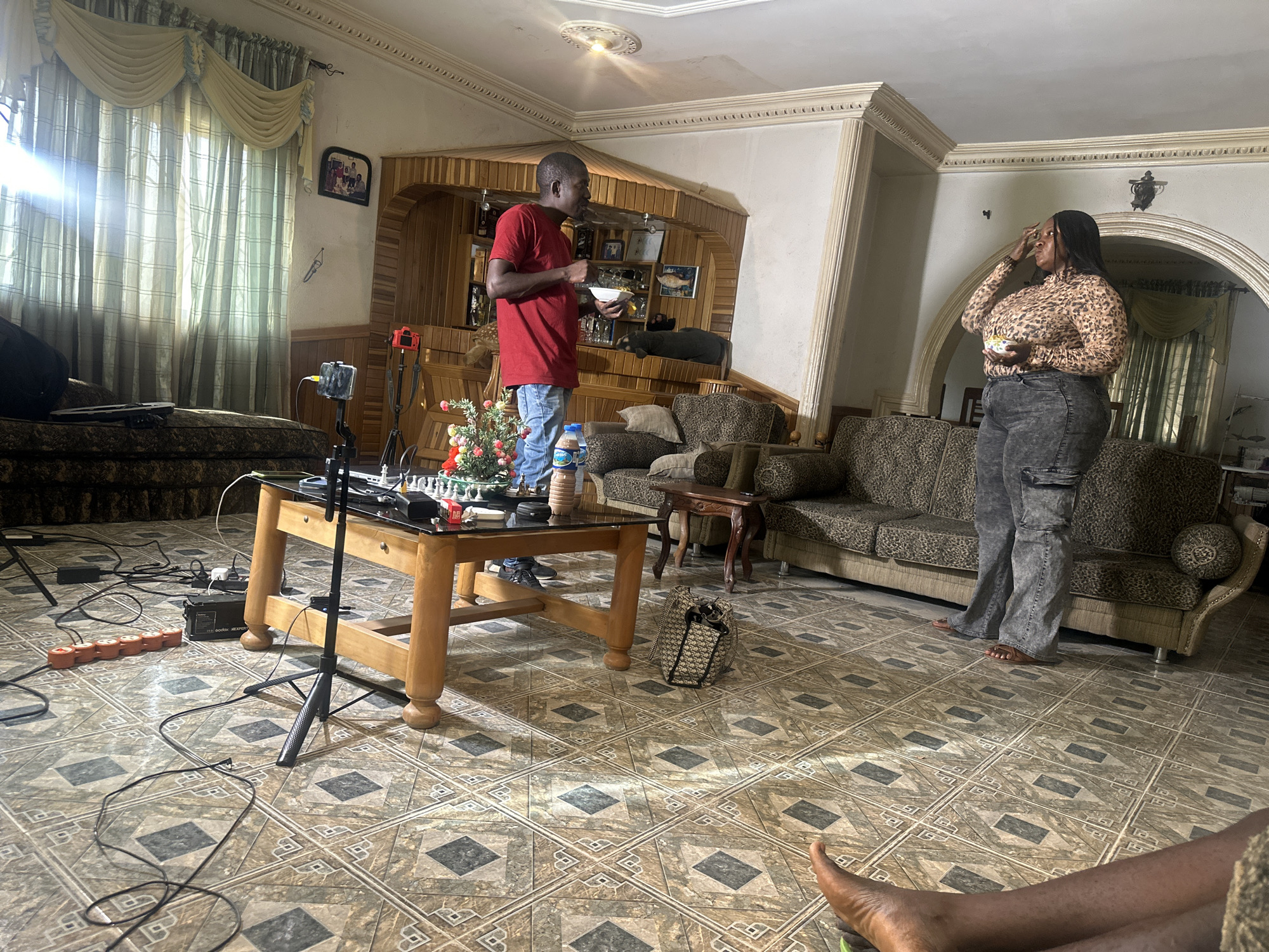 A medium-shot indoor scene captures a room with two people, a man and a woman, possibly engaged in filming a scene or creating content. The man, wearing a red t-shirt and blue jeans, is standing near a wooden coffee table and holding a bowl, possibly containing food. The woman, in a leopard-print top and gray pants, stands further away near a couch with a leopard-print pattern. Both individuals are holding small white bowls with unknown contents.
A smartphone on a tripod is aimed towards them, indicating a recording setup. A black power strip and assorted cables are sprawled across the tiled floor. The room is furnished with a couch, coffee table, and a built-in wooden display unit in the background. The floor is covered in patterned tiles. There is another couch behind the coffee table. The room is well lit from windows on the left, with curtains visible. The location appears to be a home in Jos, Nigeria.