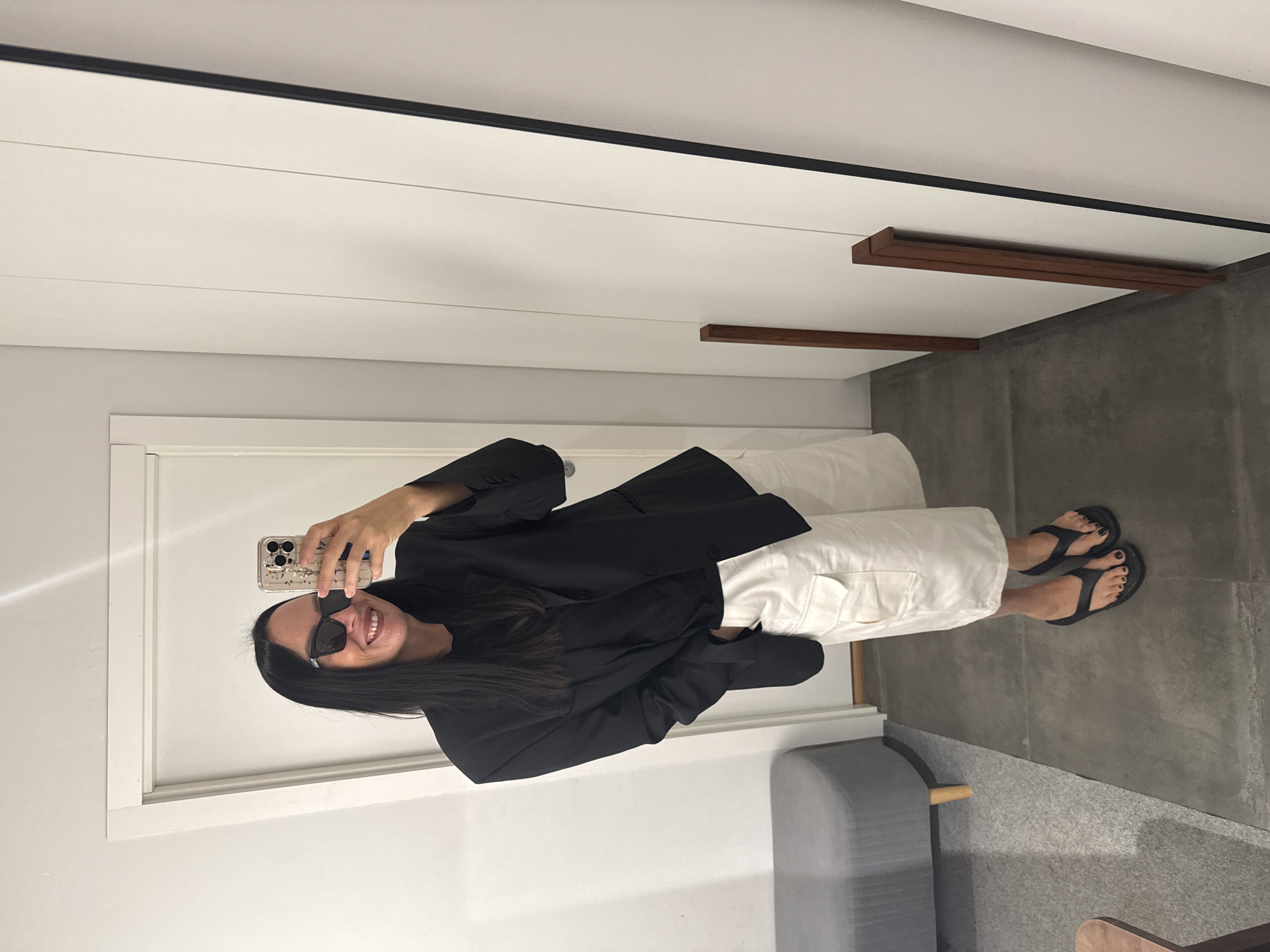 A woman is taking a mirror selfie in what appears to be a fitting room or closet. She has long dark hair, sunglasses, and is smiling. She is wearing a black blazer over a black top and white cargo shorts. She is also wearing black flip-flops. In the background, there is a gray upholstered bench, a white door, and a large mirror. The floor is tiled. There is no indication of the city of Chaikovsky, Russia.