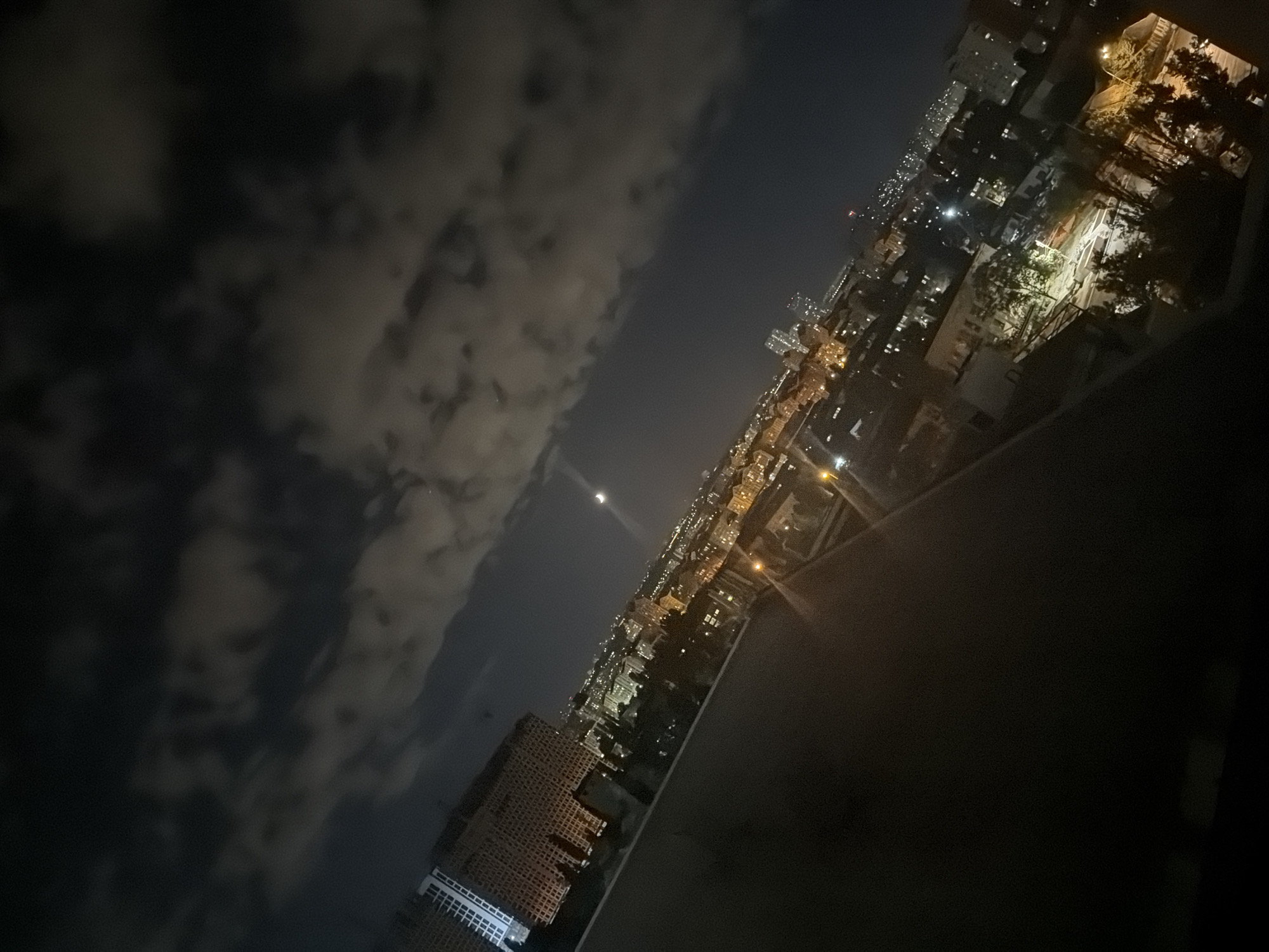The image displays an aerial nighttime view of a city, presumably Kyiv, Ukraine, from an elevated vantage point. A large, rectangular building with visible window lights is prominent on the left side of the frame. The cityscape below is illuminated by numerous lights from buildings and streetlights, creating a textured urban landscape. A crescent moon is faintly visible in the dark, cloudy sky. The foreground features a dark, slanted surface, possibly a balcony or rooftop. The overall scene conveys a sense of urban life continuing after dark.