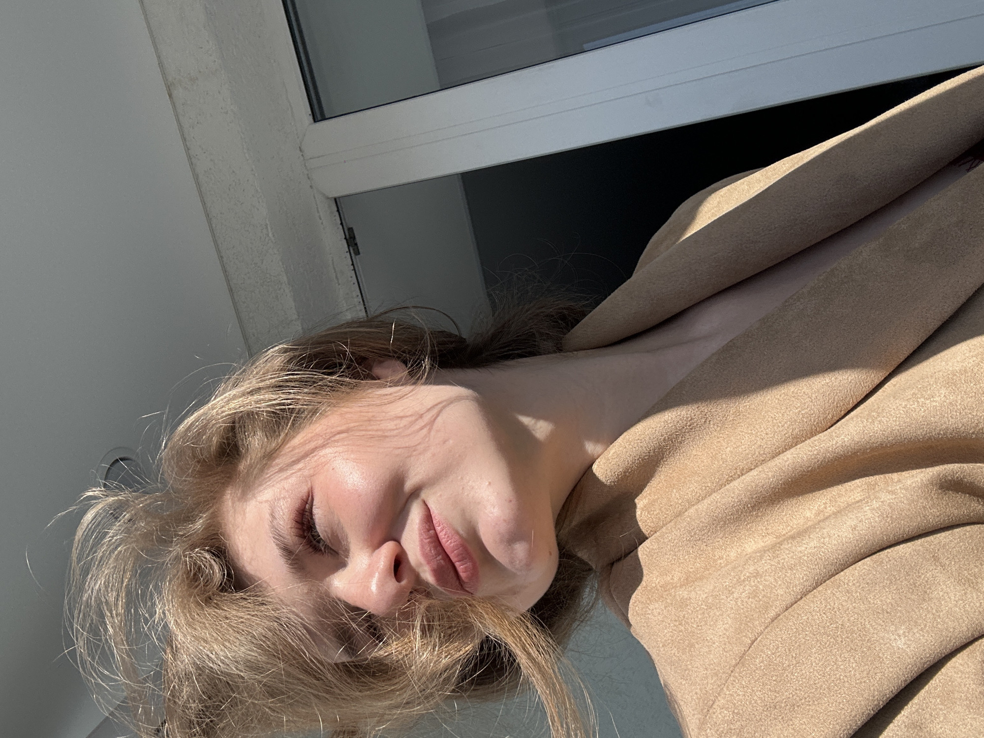 A young woman with light skin, blonde hair, and closed eyes is positioned at an angle in front of a window. Her face is partly illuminated by direct sunlight, and she's wearing a tan-colored jacket or coat. The location is likely indoors, suggested by the window and wall visible in the background. The presence of a standard white-framed window and the architecture shown is not enough to determine the location of Kazan, Russia.