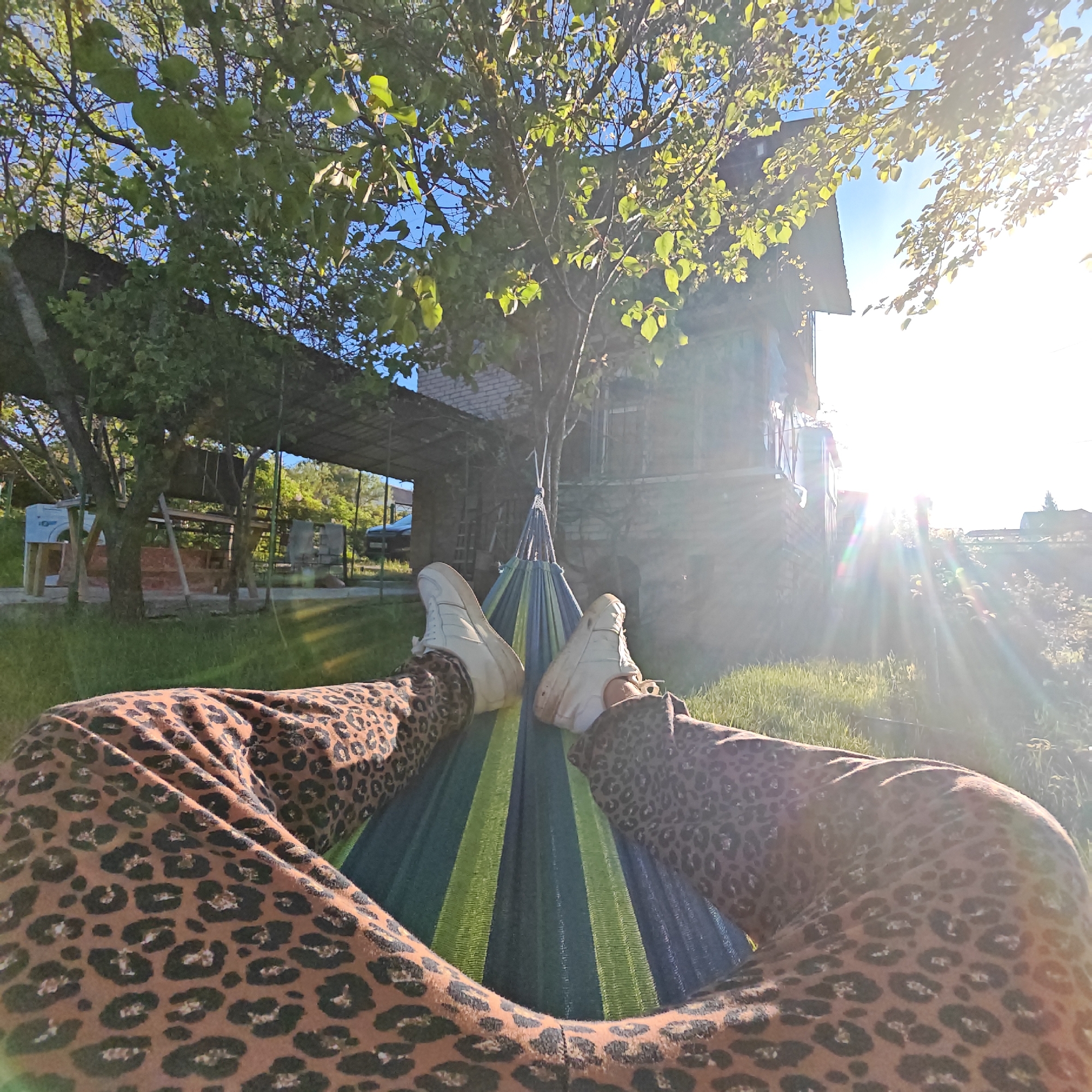 A person wearing leopard-print pants and white shoes is relaxing in a hammock in a yard. The hammock is made of blue and green stripes. In the background, there is a house with a covered porch area. Trees are visible around the house, and sunlight is shining brightly in the distance. The location is Unknown, Unknown.