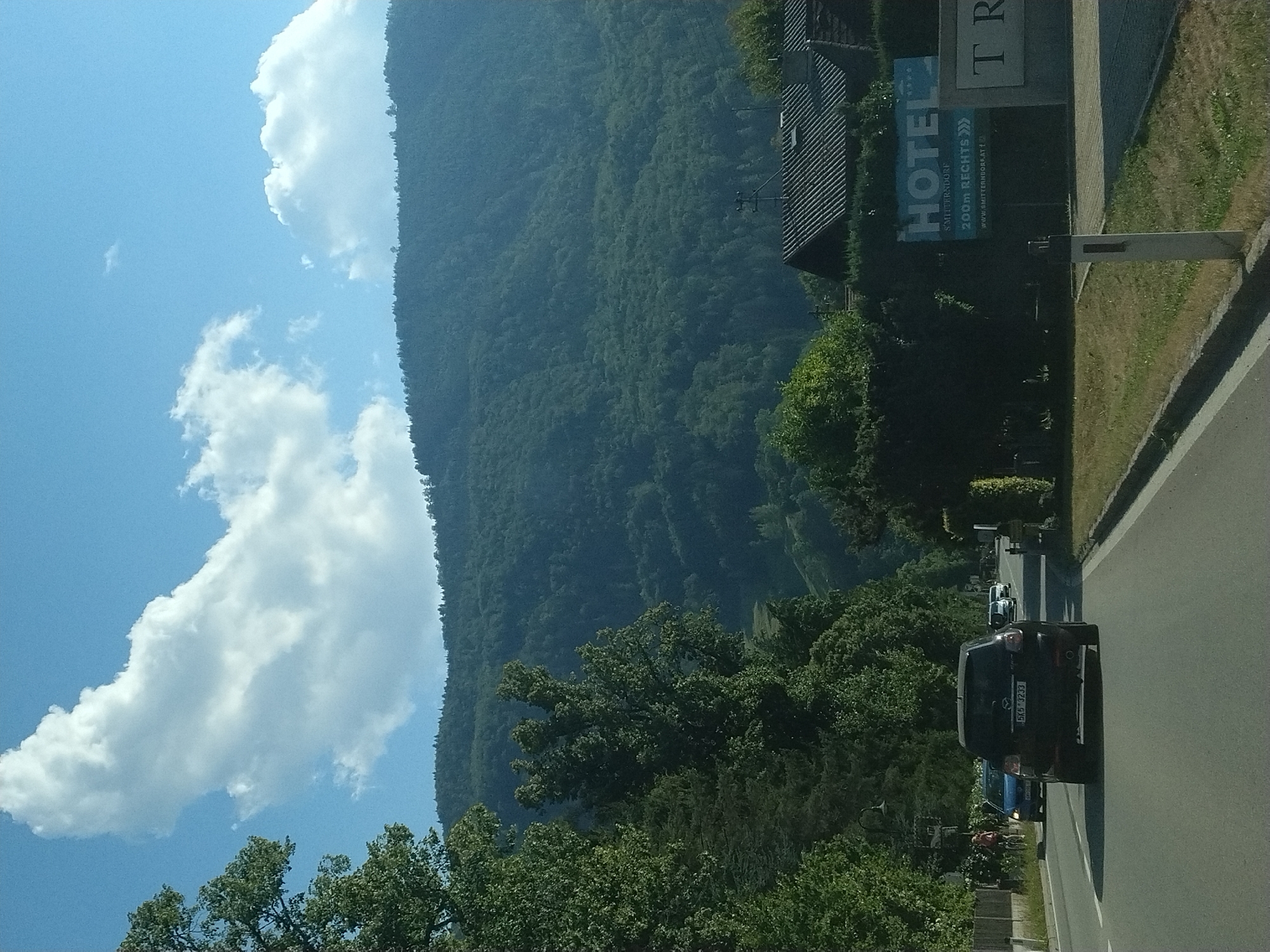 The image depicts a view from a car traveling on a road in Traunkirchen, Austria. The road curves to the left, with a dark-colored SUV in the foreground and other vehicles further ahead. Trees line the left side of the road, and a mountainous, forested landscape dominates the background under a partly cloudy blue sky. On the right side of the road, a sign for "HOTEL SMITTERNDORF" is visible, indicating proximity to the hotel, with an arrow pointing to the right and text reading "200m RECHTS". Further down the road on the right, part of a building with a sign that says "TR" is visible. A person is visible on a bicycle near the left side of the road. The overall scene is one of a scenic drive through a semi-urban or rural area with a focus on the natural landscape and nearby amenities.