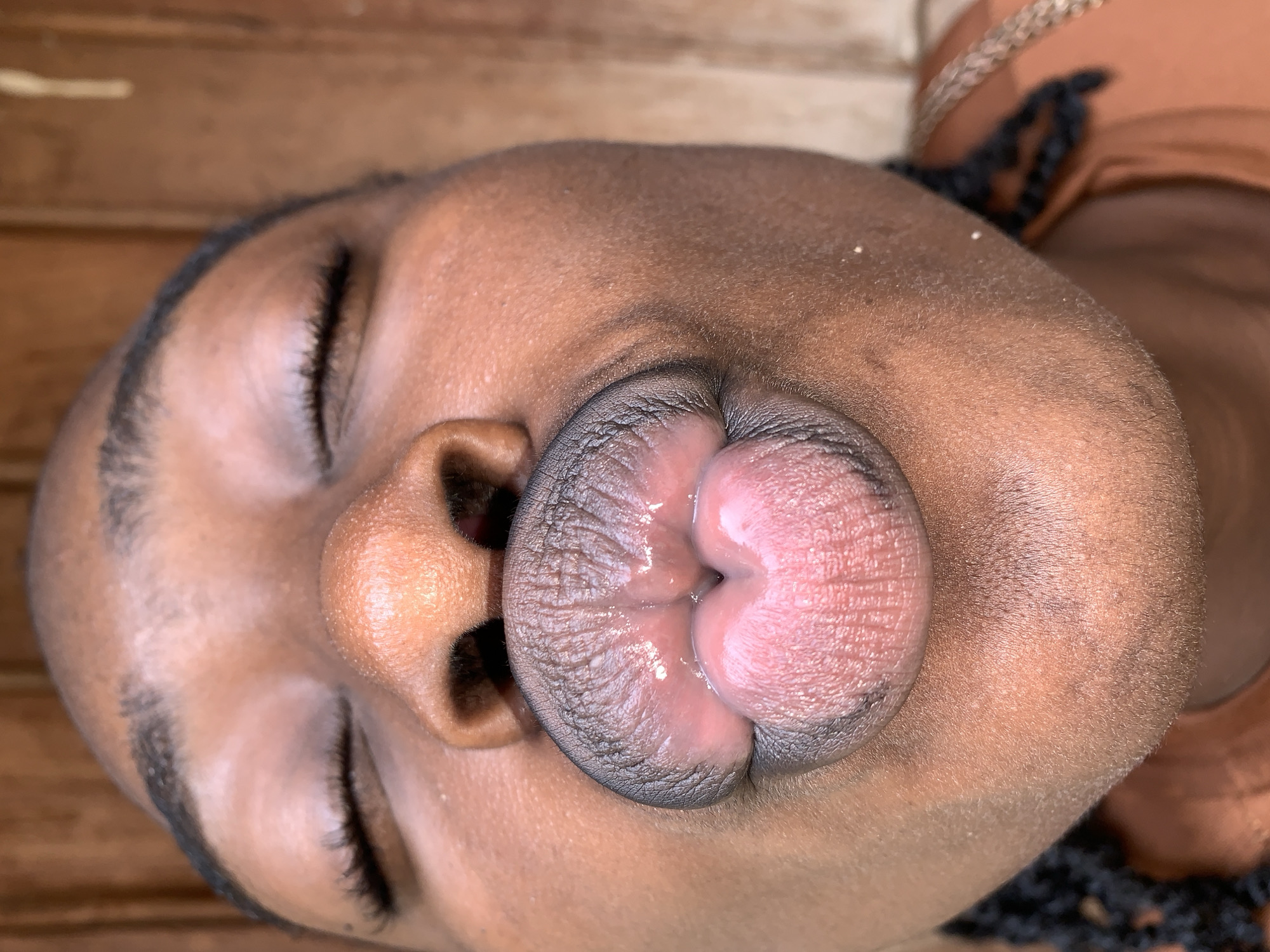 A close-up vertical portrait captures the face of an individual with dark skin, reportedly situated in the city of Rafin Par, Nigeria. The subject's eyes are closed, and their lips are prominently pursed into an exaggerated "kiss" shape, forming a distinct heart-like indentation at the center. The upper lip appears a darker, somewhat bluish-grey hue, while the interior and lower lip are a moist, lighter pinkish-red, exhibiting fine lines and texture from the pucker. Visible features include neatly groomed, dark eyebrows, closed eyelids with dark eyelashes, a broad nose with discernible pores, and smooth cheeks. Braided dark hair is visible on the left side of the frame near the chin, and a portion of a light brown or orange garment and a braided strap are seen on the lower right. The background consists of vertically aligned wooden planks, heavily blurred, suggesting a shallow depth of field. Soft, ambient lighting illuminates the subject from the front.