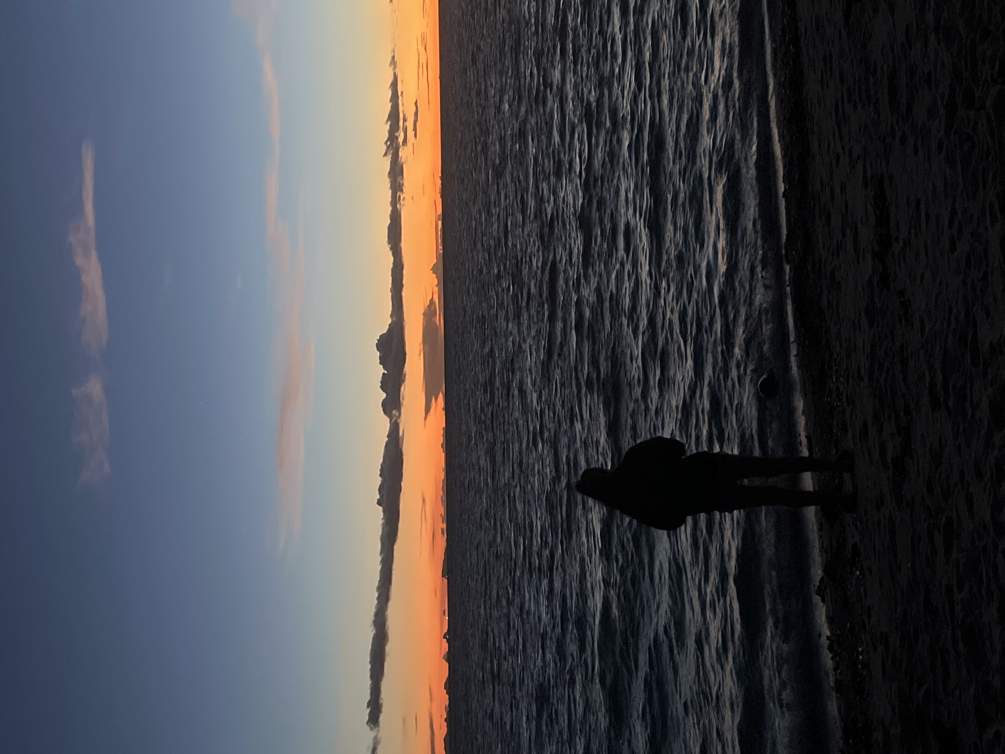 The image depicts a solitary figure silhouetted against a dusky sky and the dark, textured surface of the ocean. The sky transitions from a pale blue at the top to hues of orange and yellow near the horizon, indicating either sunrise or sunset. Thin, wispy clouds are scattered across the sky, catching the fading light. The ocean's surface appears choppy, with visible ripples and waves creating a dynamic, textured pattern.
The figure, standing on what appears to be a dark, sandy shore, is facing away from the camera, towards the water and the horizon. Their posture is relaxed, suggesting a moment of quiet contemplation or observation. The overall mood is serene and introspective, amplified by the muted colors and the vastness of the seascape. The lighting suggests it is dusk or dawn. The location context provided, ตำบลเกาะพะงัน, Thailand, suggests a tropical coastal setting. No text is visible in the image.