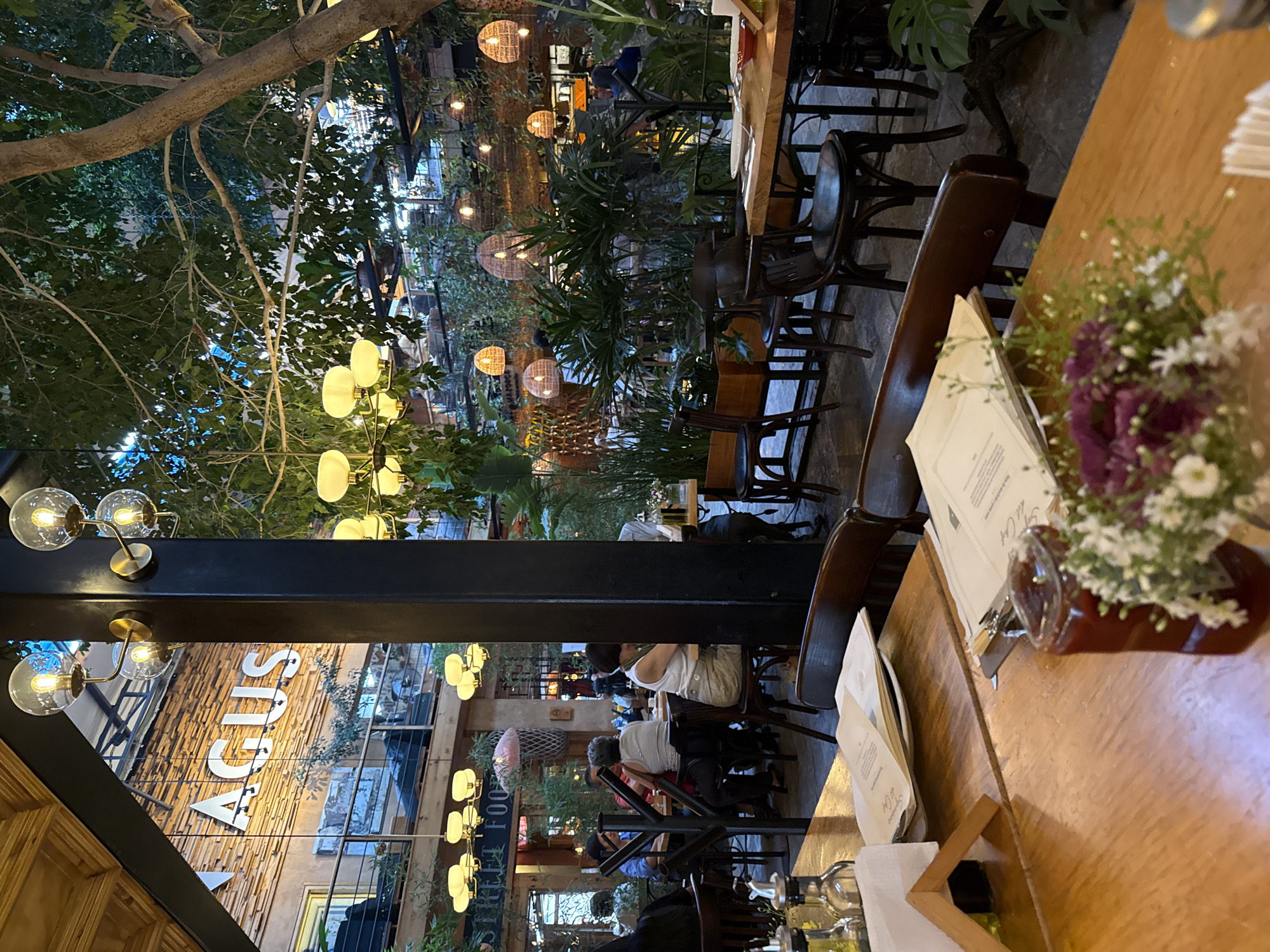 The image depicts an interior view of a restaurant, possibly located in San Cristóbal, Mexico, based on architectural and cultural context clues. Diners are seated at wooden tables and chairs within a partially outdoor setting, under trees and decorative lighting elements, including round rattan-like lanterns. A sign reads "AGUS" on a building visible in the background. A flower arrangement, menus, and condiments are present on the foreground table.