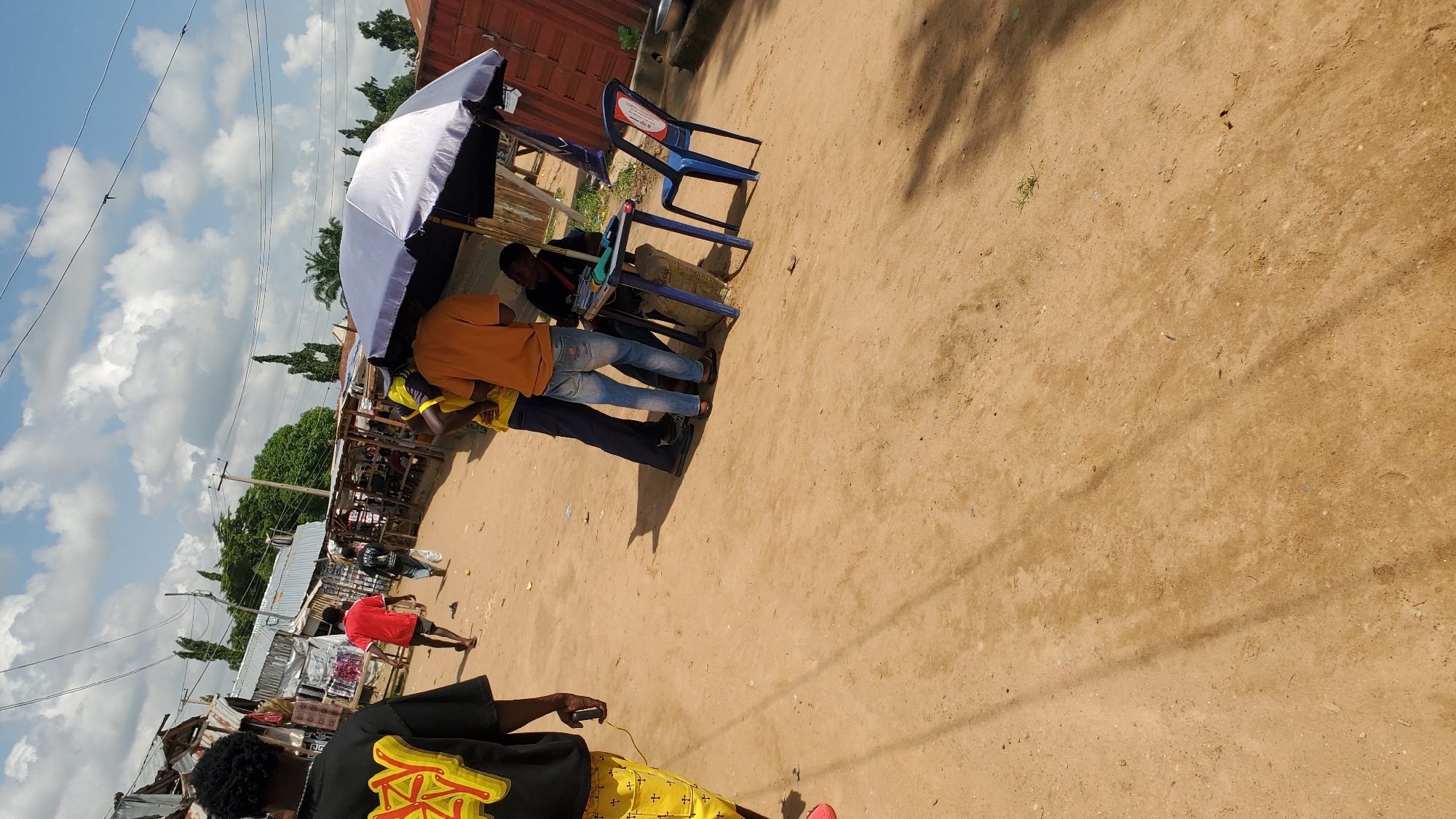 This is a medium shot, eye-level photograph taken in a dusty, outdoor market setting in Jalingo, Nigeria. The scene captures a moment of interaction around a makeshift stall. The ground is sandy and uneven, with tire tracks and scattered debris. In the foreground, a person wearing a black t-shirt with a yellow graphic design and yellow pants with a cross pattern is walking away from the camera, their left hand holding a cable. To the left of the frame, a person in a red t-shirt and dark shorts walks towards the center of the image.
The main focus is a stall set up under a large, blue and white umbrella. A blue plastic table is positioned under the umbrella, with a man in an orange t-shirt and blue jeans standing behind it, looking down. To his left, another person in a yellow t-shirt and dark pants is also standing. A blue plastic chair with a red logo on it is placed to the right of the table. A person wearing a black t-shirt is seated behind the table, appearing to be engaged in a transaction or discussion with the person in the orange t-shirt. There are some items on the table, including what appears to be a microphone and some cables. In the background, there are several market stalls with corrugated metal roofs and wooden structures, as well as trees and utility poles. The sky is partly cloudy with a bright sun. The overall impression is a busy, informal marketplace.