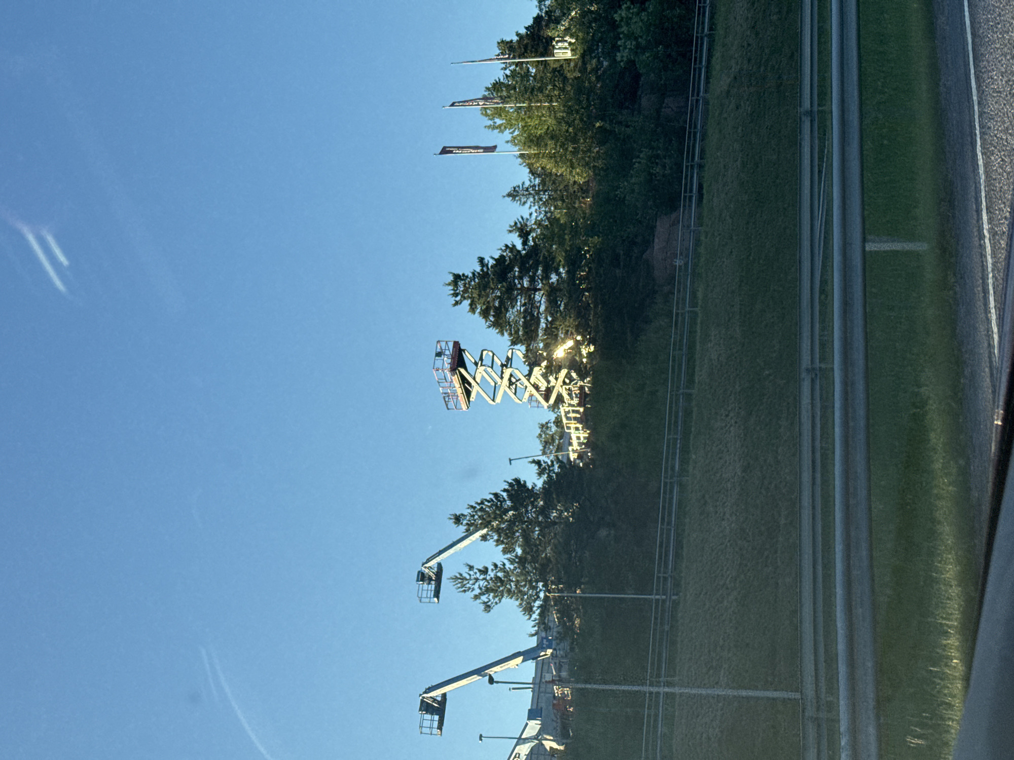 The image depicts an outdoor scene under a clear blue sky. In the background, there is a treeline on a grassy hill. Various construction vehicles, including two boom lifts and a scissor lift, are visible among the trees. Three dark flags with white lettering are planted on the hill. The foreground shows a highway with guardrails and a grassy embankment. The overall impression is that of a construction site or an area undergoing development, possibly near a road.