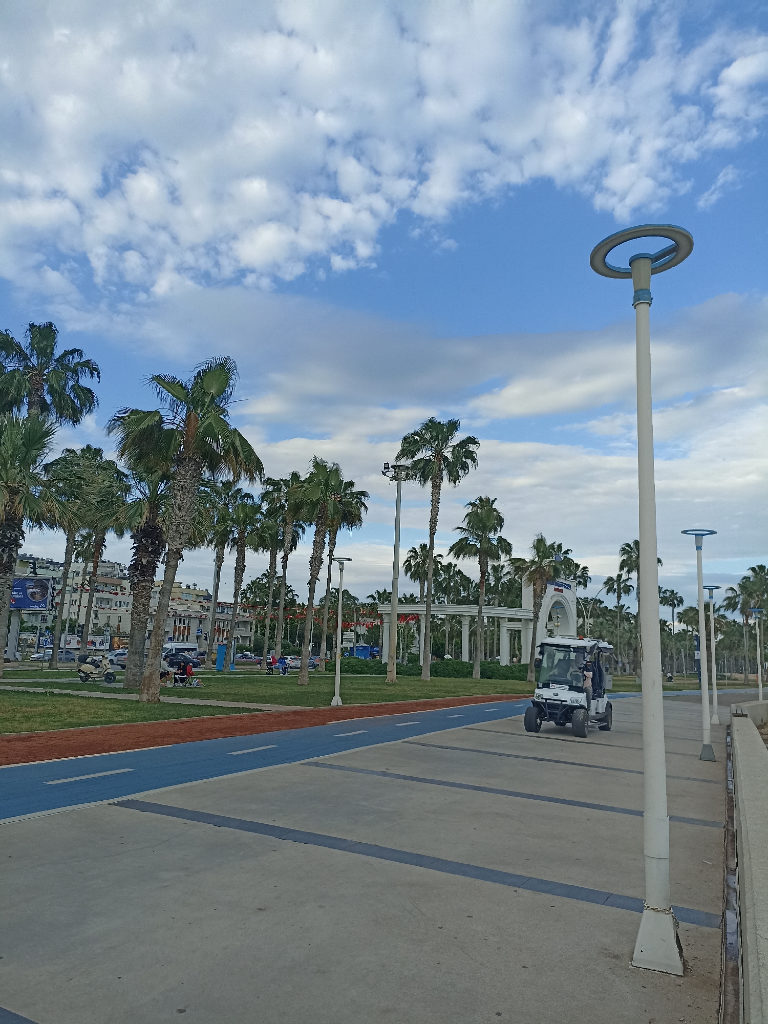 The scene is a public promenade in Yenişehir, Turkey, featuring a concrete walkway marked with blue lines, adjacent to a red running track and grassy area dotted with palm trees. The sky is partly cloudy. A white electric vehicle with two visible occupants is moving along the walkway. Tall, white lamp posts with circular light fixtures line the edge of the promenade. In the background, an arched entrance structure is visible, surrounded by more palm trees. Beyond the trees are buildings and signs of urban activity.