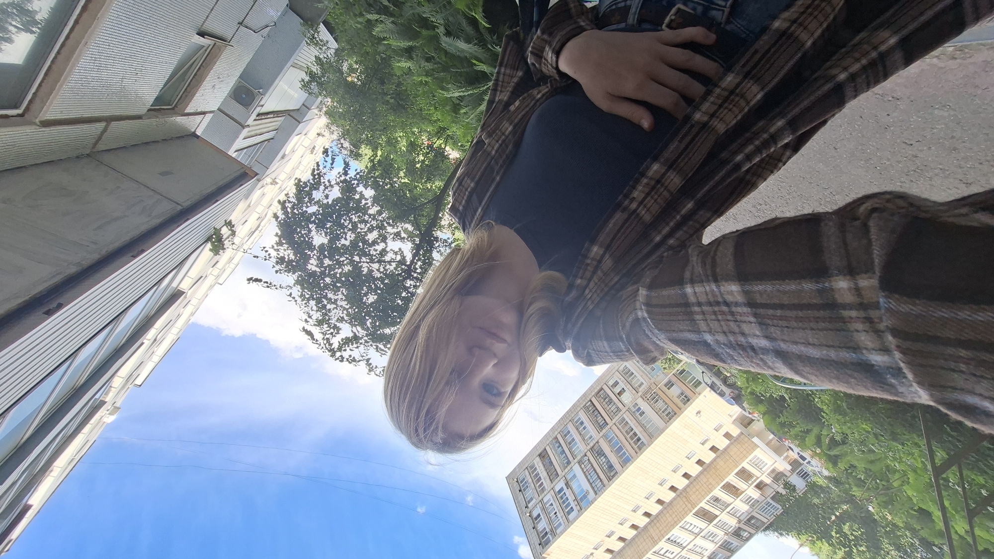 A low-angle, wide shot captures a person taking a selfie in an urban outdoor setting. The person, with blonde hair and fair skin, is wearing a dark top and a brown and beige plaid shirt. Their face is partially obscured by their hair and hand, and they appear to be looking down at the camera.
The background features a clear blue sky with some wispy clouds. To the left, a modern, multi-story building with a metallic facade and visible windows is prominently featured. On the right, another taller apartment building with numerous windows can be seen, interspersed with lush green trees and foliage. The ground is a paved surface, likely asphalt, suggesting a walkway or street.
The image appears to be taken during daylight hours, as indicated by the bright sky and the natural lighting. The overall atmosphere is casual and relaxed. The orientation of the buildings and trees suggests the photo was taken in a residential area. No visible text is present in the image. The perspective of the selfie, looking upwards towards the person and the sky, is a common photographic style for self-portraits. The scene is likely a street or courtyard in Chaikovsky, Russia, given the architectural style of the buildings.