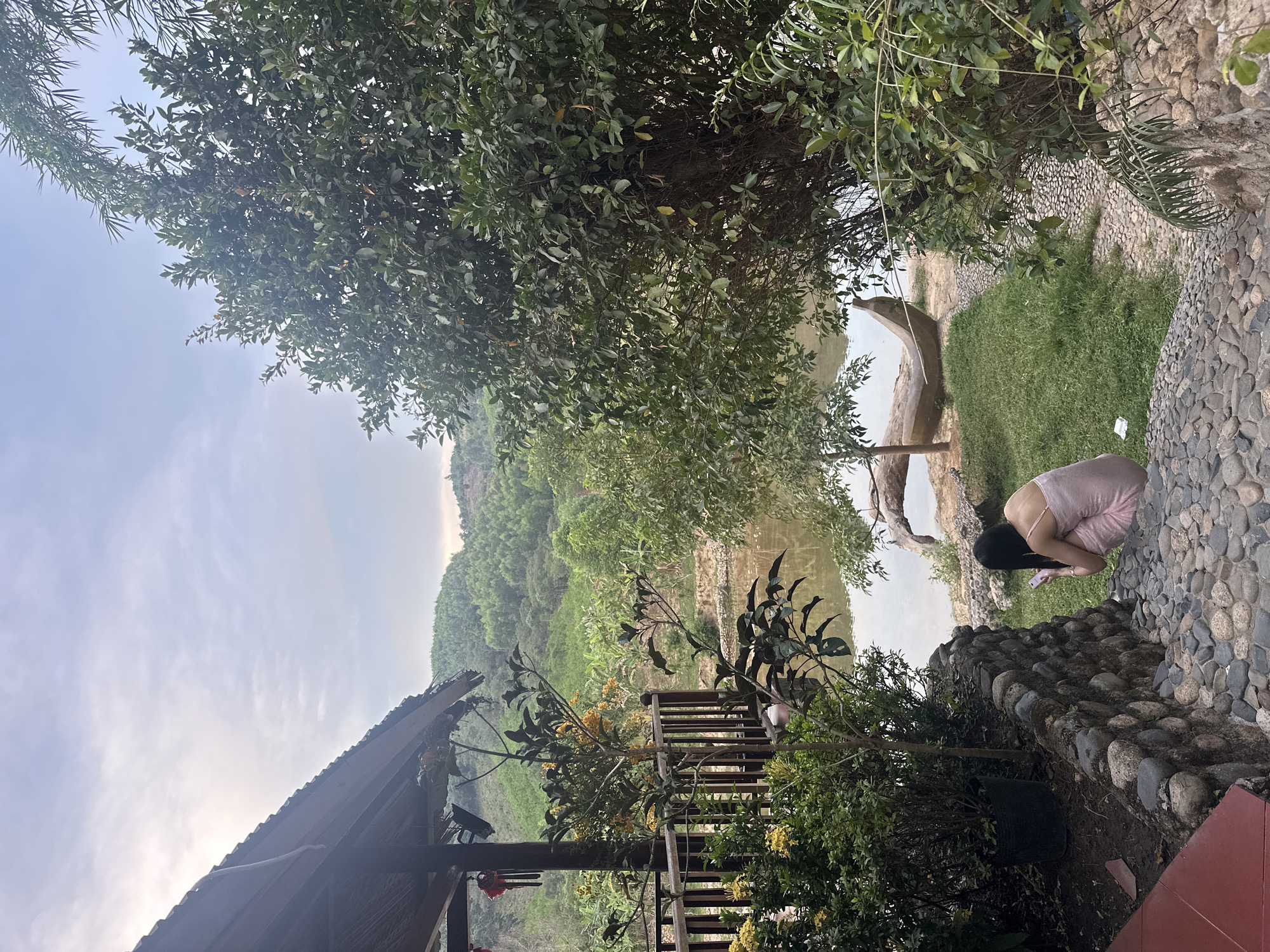 The image shows an outdoor scene in Nam Khanh Vinh, Vietnam. A woman in a light pink dress is crouched down near a stone wall, seemingly looking at her phone. A river or pond is visible, reflecting trees and sky. A wooden structure, possibly a deck or gazebo, is partially visible in the lower left corner. Lush greenery, including trees and various plants, dominates the landscape. The sky is partly cloudy.