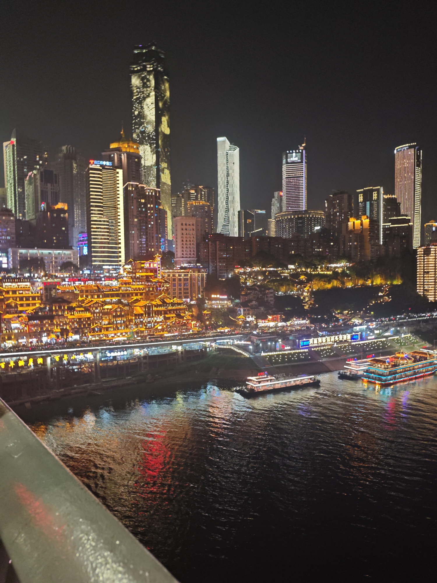The image shows a night view of the Hongyadong historic area in Yuzhong District, Chongqing, China. The buildings are illuminated with yellow lights, and there are people on the walkways. In the foreground, there is a body of water with reflections of the city lights and boats traveling along the river. The background features multiple skyscrapers. A railing is visible in the very bottom left corner.