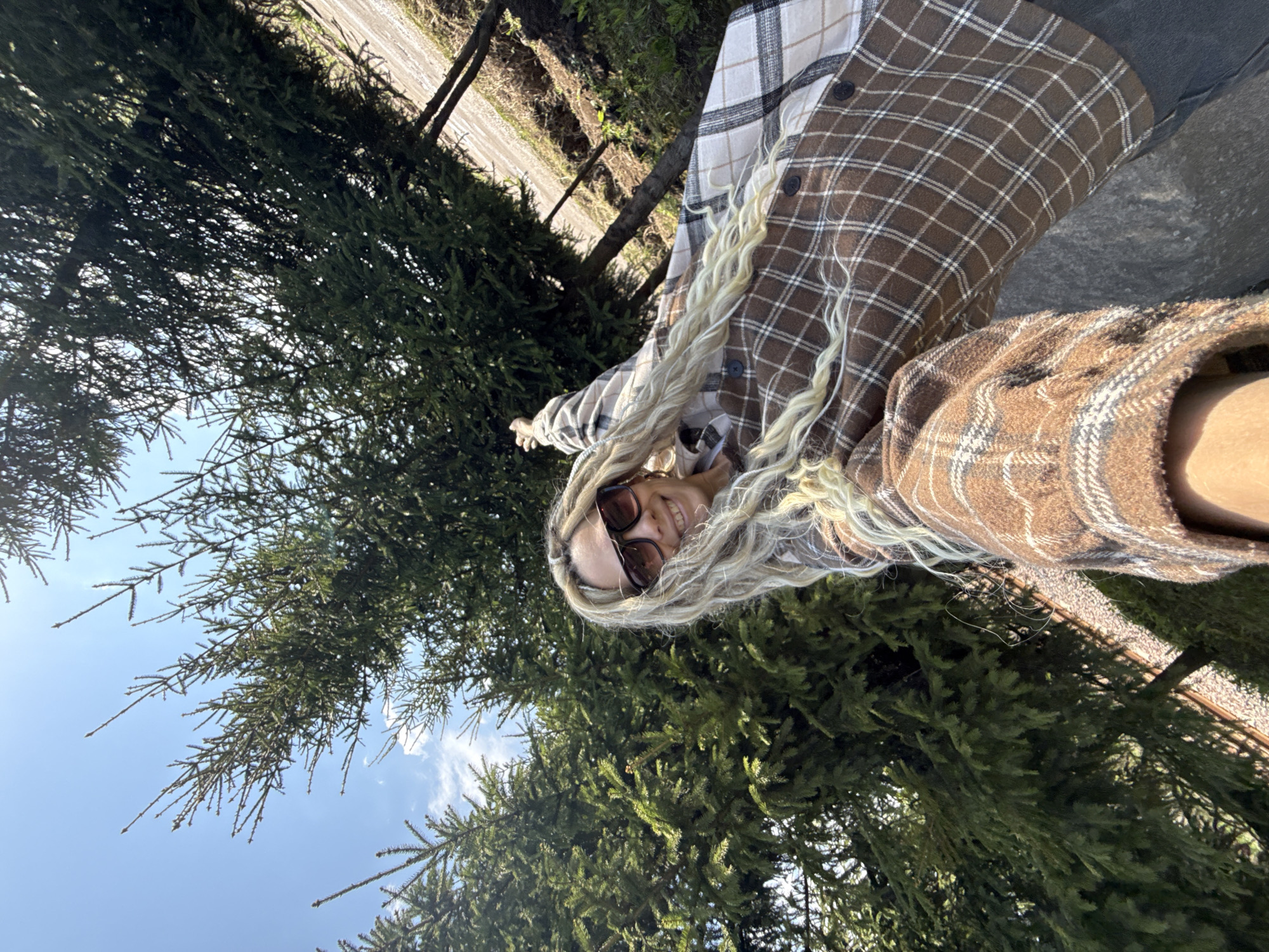 The image shows a woman with blonde dreadlocks wearing sunglasses and a brown and beige plaid shirt. She is standing outdoors with evergreen trees in the background. The sky is visible in the upper left corner. Based on the image content, the exact location within Rusanovo, Russia, cannot be determined.