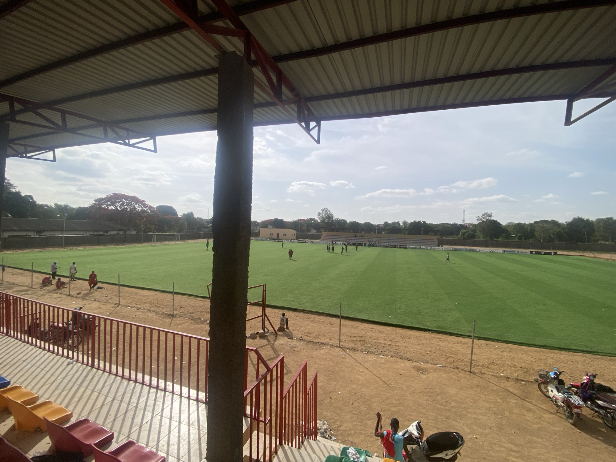 The image depicts a stadium in Tudun Wada, Nigeria. The stadium has a green soccer field where a group of people are gathered, presumably players in practice or a game. A rusty corrugated metal roof covers part of the stadium, supported by red metal beams and a large concrete pillar in the foreground. The stadium seating is sparse, consisting of various colored plastic chairs. Red metal railings run along the stadium's edge. Outside the railings, the ground is sandy. Several motorcycles are parked in the dirt area near the stadium. Some people are seen near the stadium, both on the field and in the seating area. The sky is partially cloudy. The photo appears to be taken from a high vantage point within the stadium.