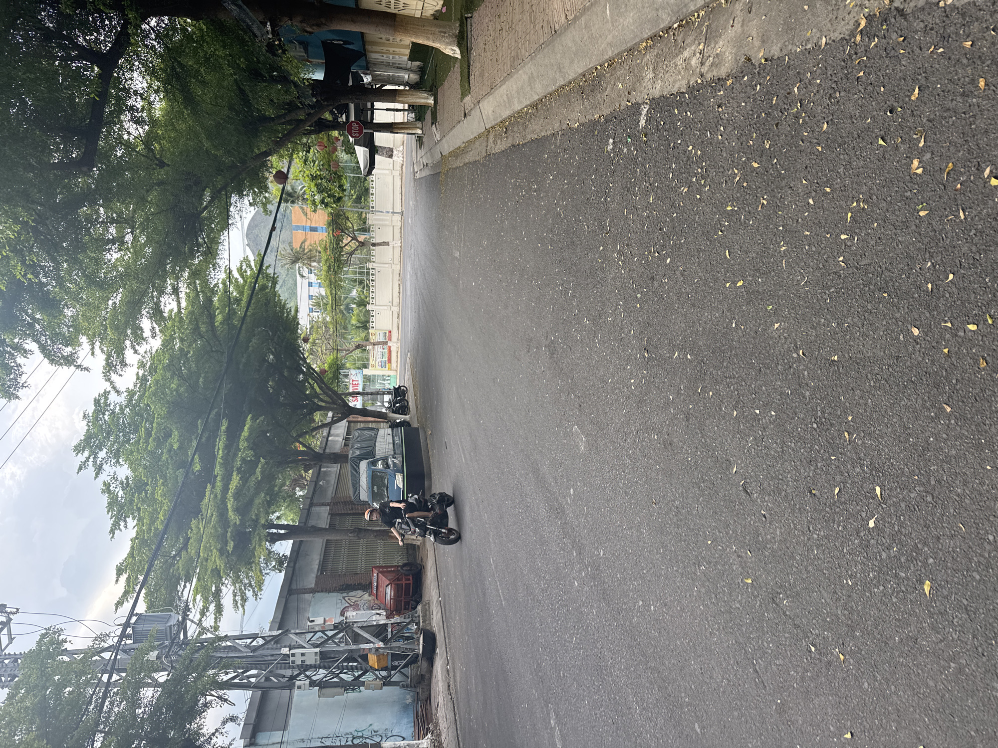 The image shows a street scene in Bac Nha Trang Ward, Vietnam. A person is riding a dark-colored motorcycle down the middle of the asphalt road. The rider is wearing a helmet and dark clothing. To the left of the road, there is a blue building with a large metal utility pole in front of it. Further down the street, a blue truck with a black tarp is parked. On the right side of the road, there is a sidewalk with red brick paving and a concrete curb. Several trees with green foliage line the street. In the background, there is a building with orange and blue accents and a mountain in the distance. A red stop sign is visible on the right side of the road. The sky is overcast. Fallen leaves are scattered on the road and sidewalk.