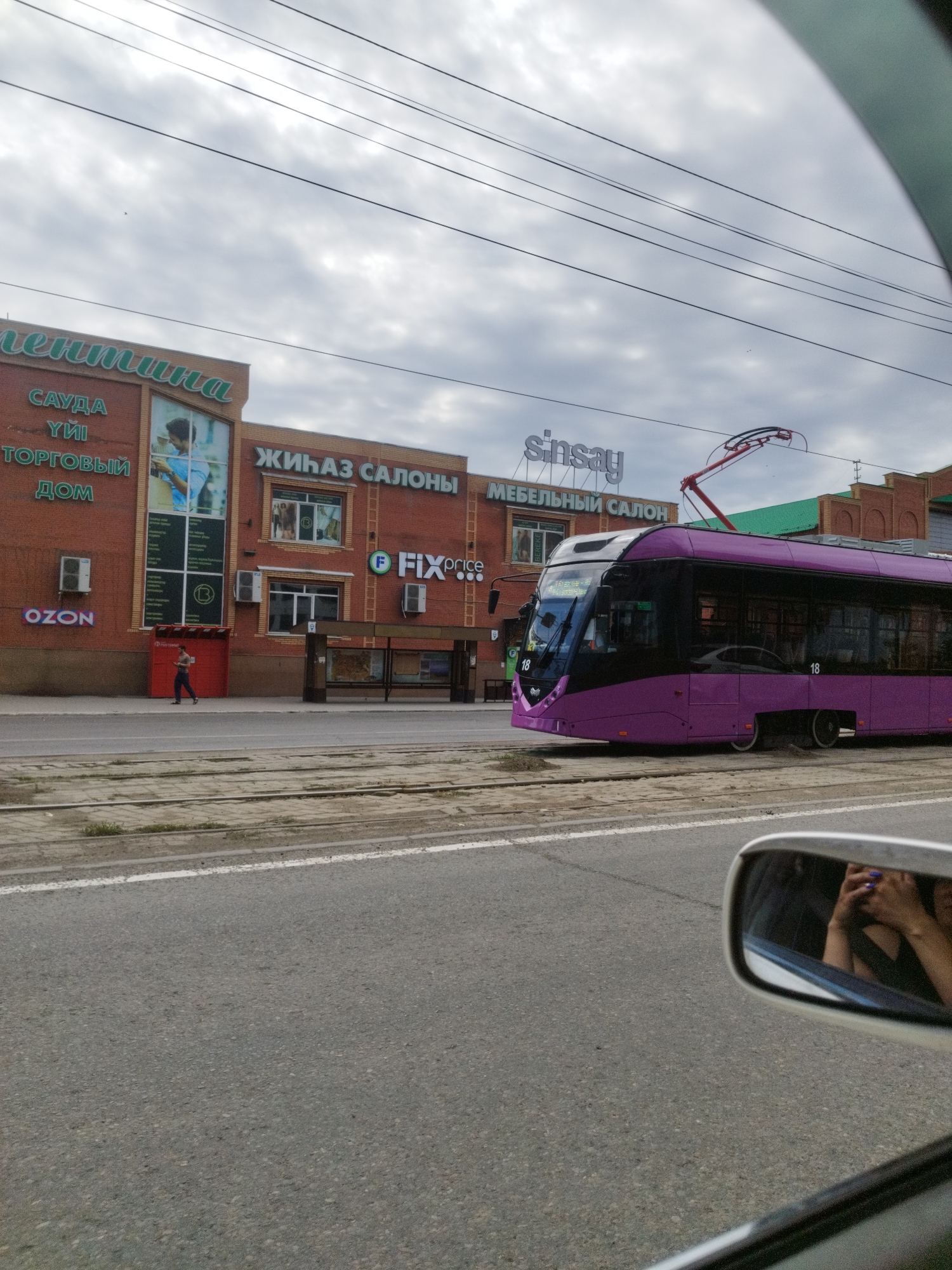 The image shows a purple tram moving along a street in Oskemen, Kazakhstan. The tram is modern with large windows and the number 18 is visible on its side. In the background, there are brick buildings with various signs, including "Sinsay," "Fix Price," "Ozon," and "Furniture Salon." A bus stop is visible on the right side of the street. The sky is cloudy, and power lines are strung across the top of the image. A person is walking on the sidewalk in front of one of the buildings. The photo is taken from a car, as a rearview mirror reflecting a person holding a phone is visible in the bottom right corner.