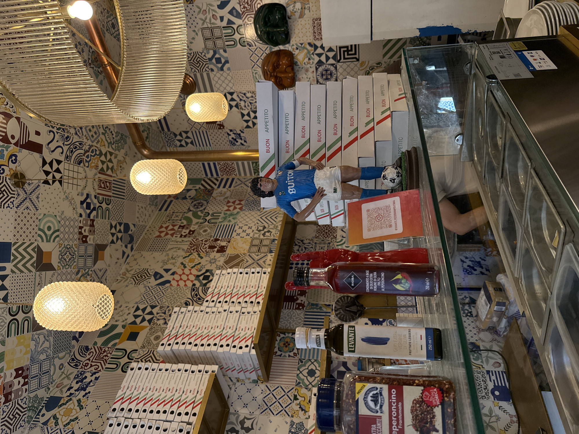The image captures the interior of a vibrant Italian eatery or pizzeria in Milan, Italy, characterized by eclectic decor and a prominent display. The walls and possibly ceiling are covered in a rich tapestry of patterned tiles featuring various geometric and traditional designs in a diverse color palette. Several ornate light fixtures illuminate the space, including a large, circular, ribbed chandelier and multiple smaller, textured spherical pendant lights.
In the foreground, a glass display counter showcases an array of Italian food products. Notable items include bottles of "LEVANTE OLIO EXTRA VERGINE DI OLIVA FRUTTATO LEGGERO" and "RIOBA CONDIMENTO A BASE DI OLIO EXTRA VERGINE DI OLIVA AL PEPERONCINO." A large container of "NOVITA Drogheria & Alimentari 1888 Peperoncino Frantumato" (crushed red pepper) is also visible. Below the counter, a reflective surface reveals part of a refrigerated prep station with metal inserts.
Dominating the midground are numerous stacks of white boxes, likely pizza boxes, all bearing the text "BUON APPETITO" and a green, white, and red striped design, reminiscent of the Italian flag. Perched atop one stack of boxes is a figurine of a soccer player, clearly identifiable as Diego Maradona in a blue jersey with "BUITON" printed on it, alongside a small soccer ball. Above the boxes, two decorative masks—one dark green and one brown—adorn a shelf. A red card with a QR code and "@TARANTE PIZZERIANAPOLETANA" provides a specific location cue. No people are directly visible, though a partial reflection in the glass suggests a person nearby. The scene is static, depicting the establishment's prepared setting rather than an active event.