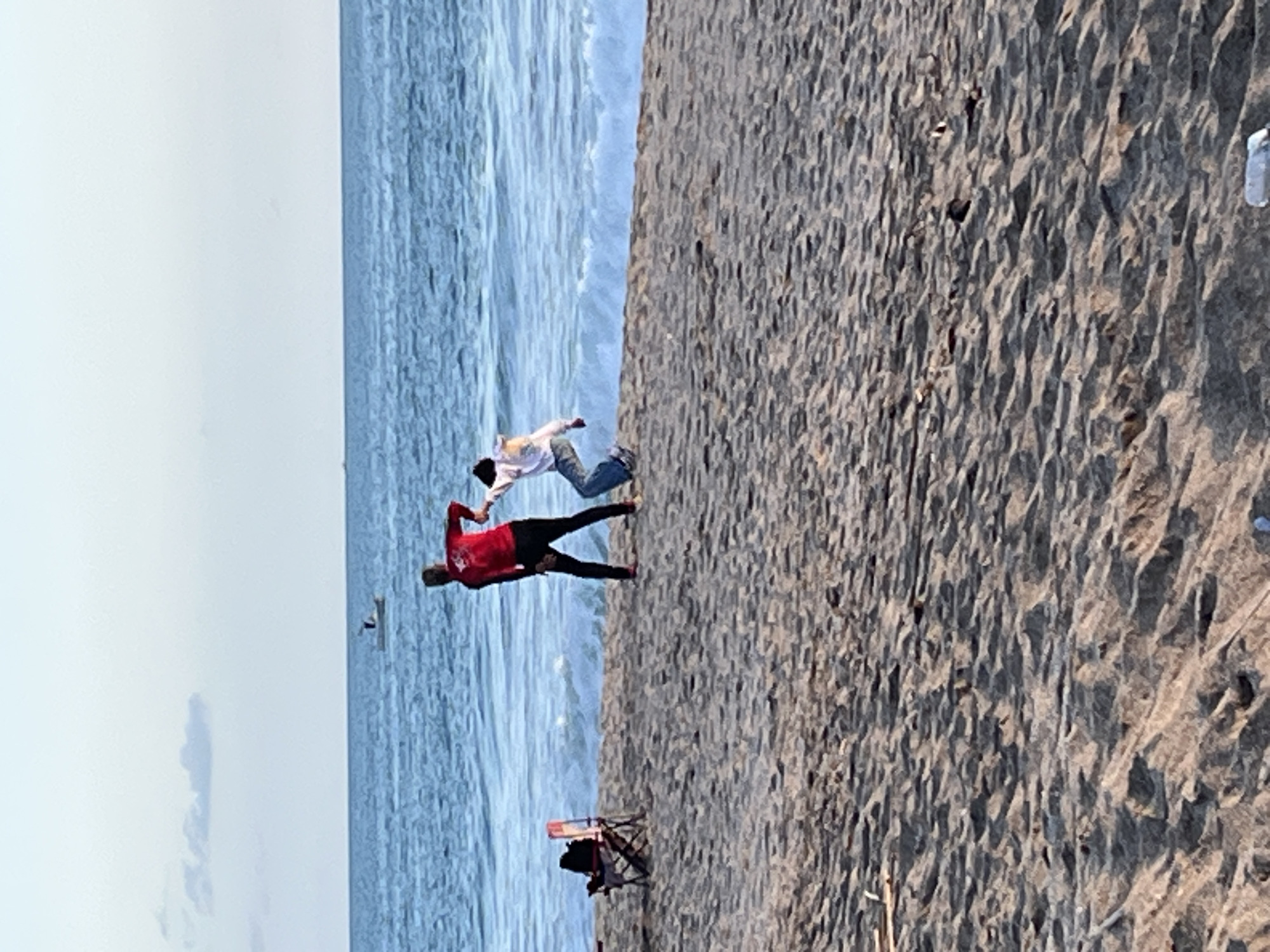 The image depicts a beach scene. Two people are near the shoreline: one wearing a red shirt and dark pants and another wearing a white shirt and jeans. They appear to be interacting. A beach chair with a person sitting in it is located further down the beach. Debris is visible on the sand. The location is most likely a beach in Alanya, Turkey, given the description provided in the prompt.