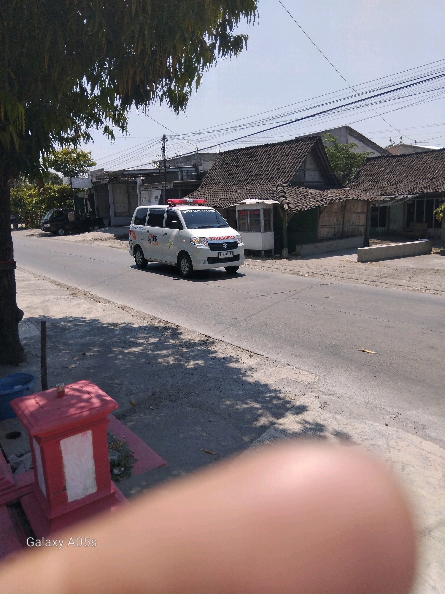 An outdoor, eye-level, medium shot shows a white ambulance with red flashing lights driving on a paved road. The ambulance has "AMBULANCE" written on its side in red lettering, as well as a logo with "BRI" in blue lettering. The ambulance also has a license plate that reads "H 9381 PG". To the left of the ambulance, a dark-colored truck is parked on the side of the road. In the foreground, a blurred finger is visible. Behind the ambulance, there are several buildings with tiled roofs, some appearing to be homes and others possibly shops or businesses. The sky is clear and blue. The environment appears to be a street in Karanganyung, Indonesia, with a sidewalk and trees lining the road.
