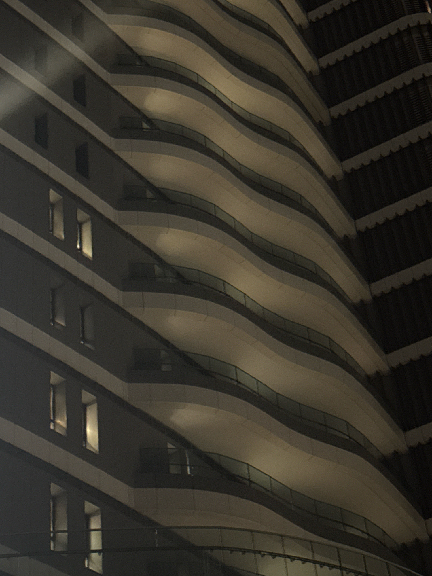 The image captures a close-up, low-angle, night-time view of a modern, curved building with staggered balconies. The building's facade is characterized by alternating bands of dark material and lighter, cream-colored curved surfaces, creating a wave-like effect. Numerous rectangular windows, some illuminated from within, are visible on the dark sections of the building. Glass railings line the edges of the balconies. The building appears to be in a developed urban area, suggested by the architectural style and the presence of other structures hinted at on the right side of the frame, which feature a darker, textured facade with a serrated edge. No people are visible in the image. The overall impression is one of contemporary urban architecture at night. This building is located in Germasogeia Municipality, Cyprus.