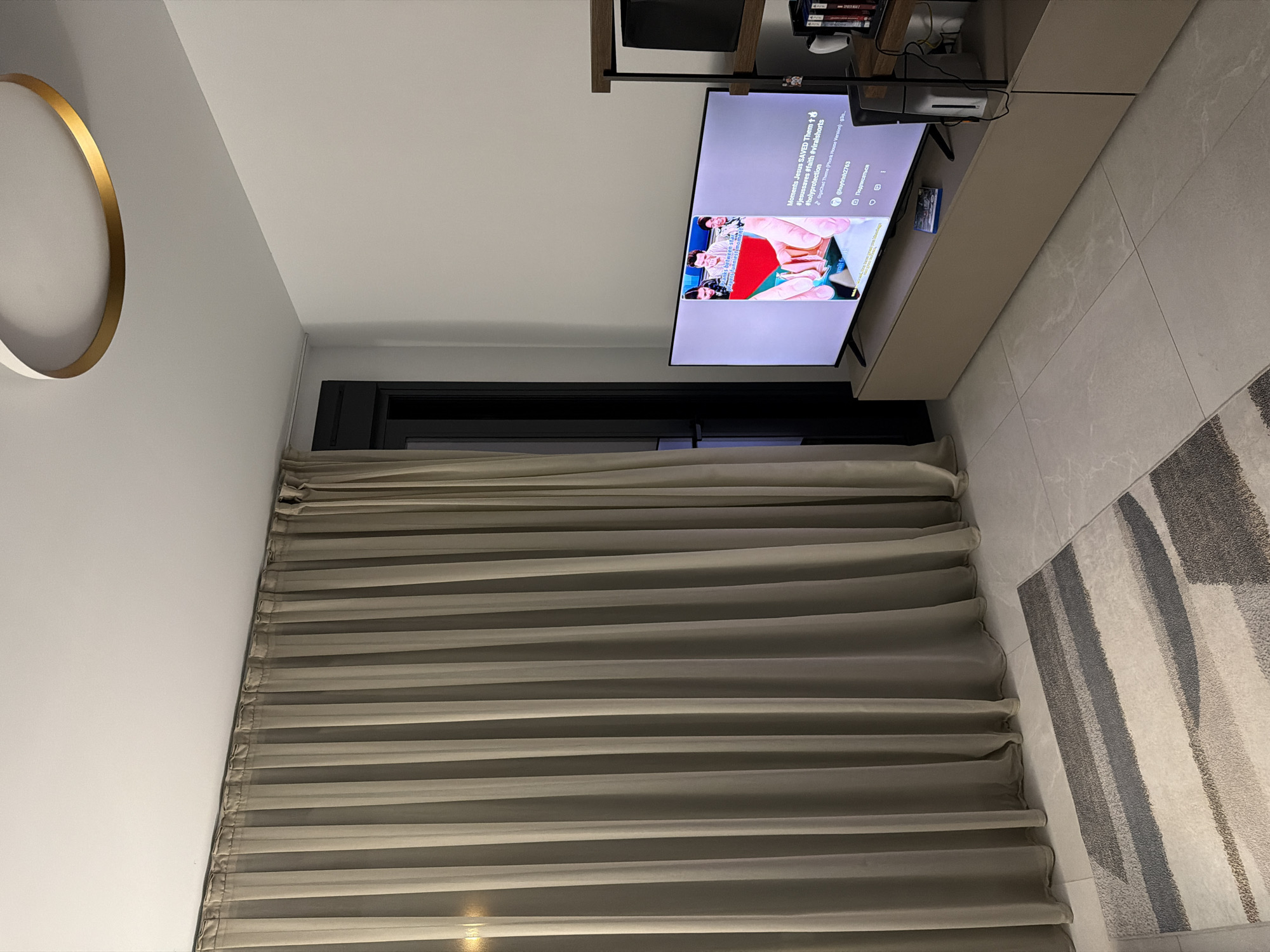 The image shows a modern living room in Finestrat, Spain. The room features a large, sheer beige curtain covering a window or sliding door on the left side. A contemporary rug with abstract grey and white stripes is placed on a light-colored tiled floor. In the center of the room, a large flat-screen television displays a video with text overlays, including "Moments Jesus SAVED Them 🙏 † #jesussaves #faith #viralshorts #holyprotection". The TV is mounted on a minimalist beige entertainment unit. To the right of the TV, a dark object, possibly a speaker, is visible on a wooden shelf unit, alongside some books and what appears to be a white mug. A circular ceiling light fixture with a gold accent is visible at the top of the image. The walls are painted white. The scene is dimly lit, suggesting it might be evening or the curtains are drawn to block natural light. No people are visible in the image.