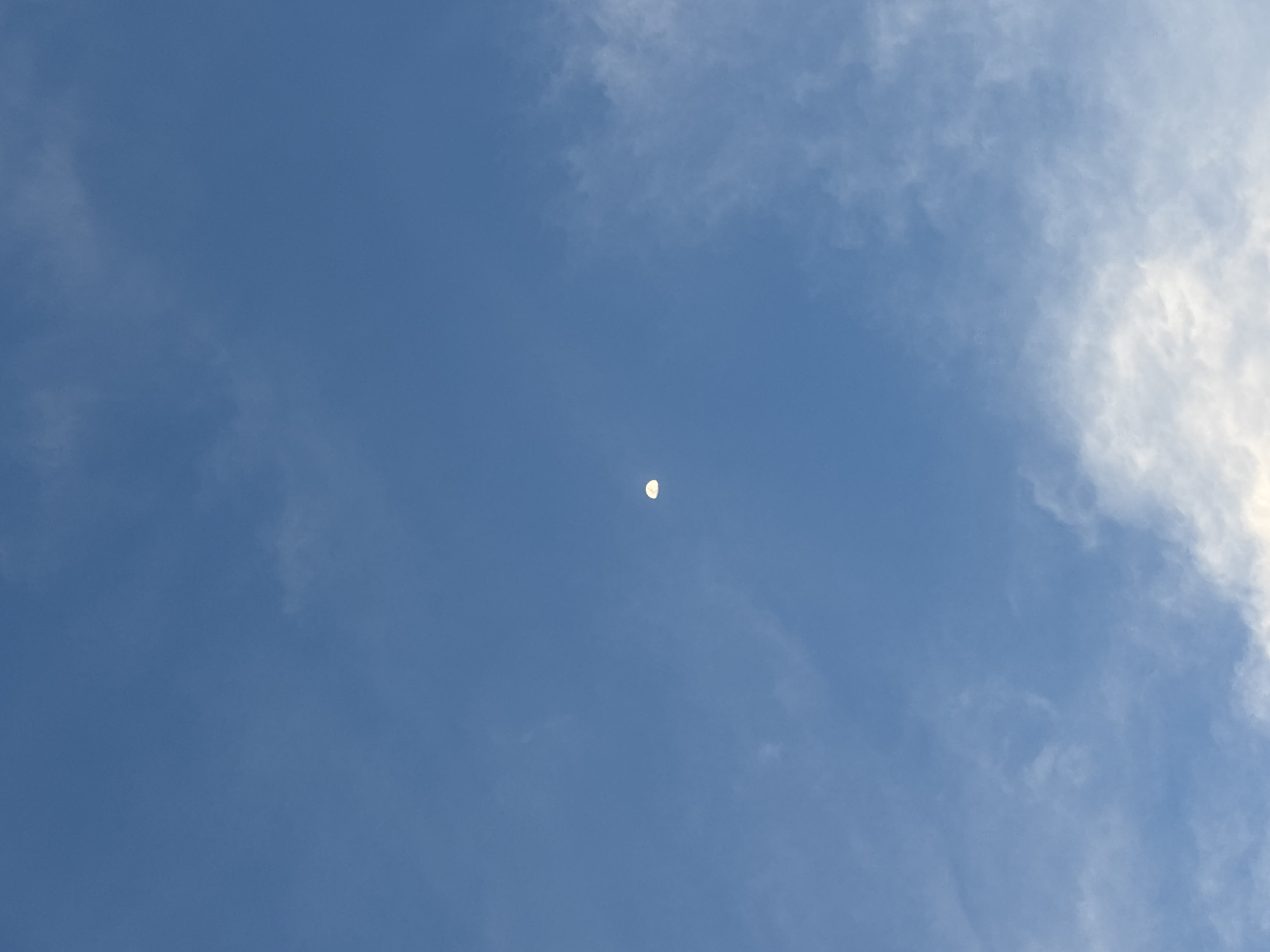 The image shows a daytime sky in Santa Clara Ocoyucan, Mexico. The sky is blue with scattered white clouds. A crescent moon is visible near the center of the frame.
