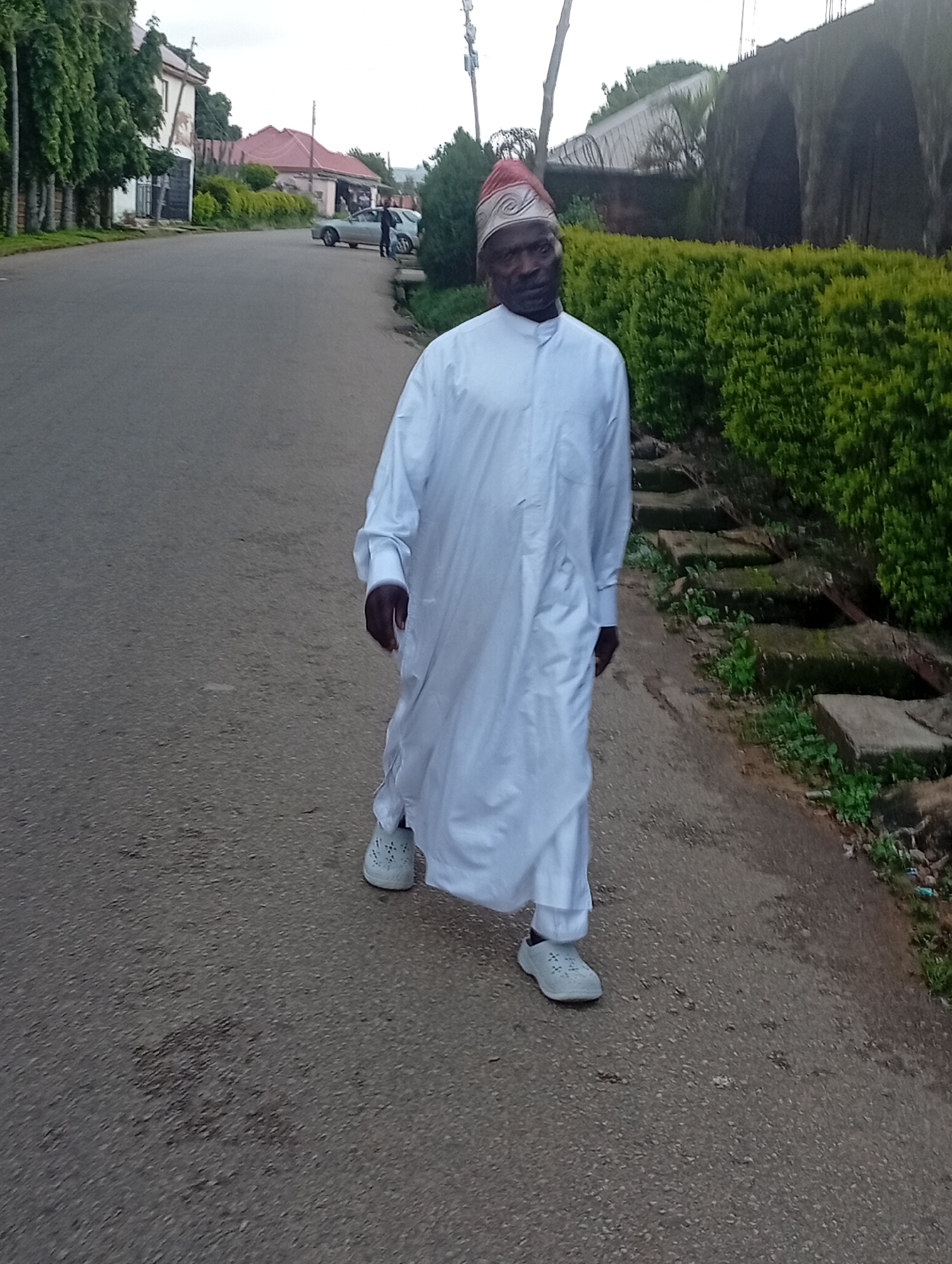 An African man wearing a white robe and a patterned hat walks on a paved road in front of a manicured hedge. The man is facing forward, with his right leg slightly ahead of his left. He has his hands by his sides. The road is lined with trees and buildings in the background. The sky is overcast.