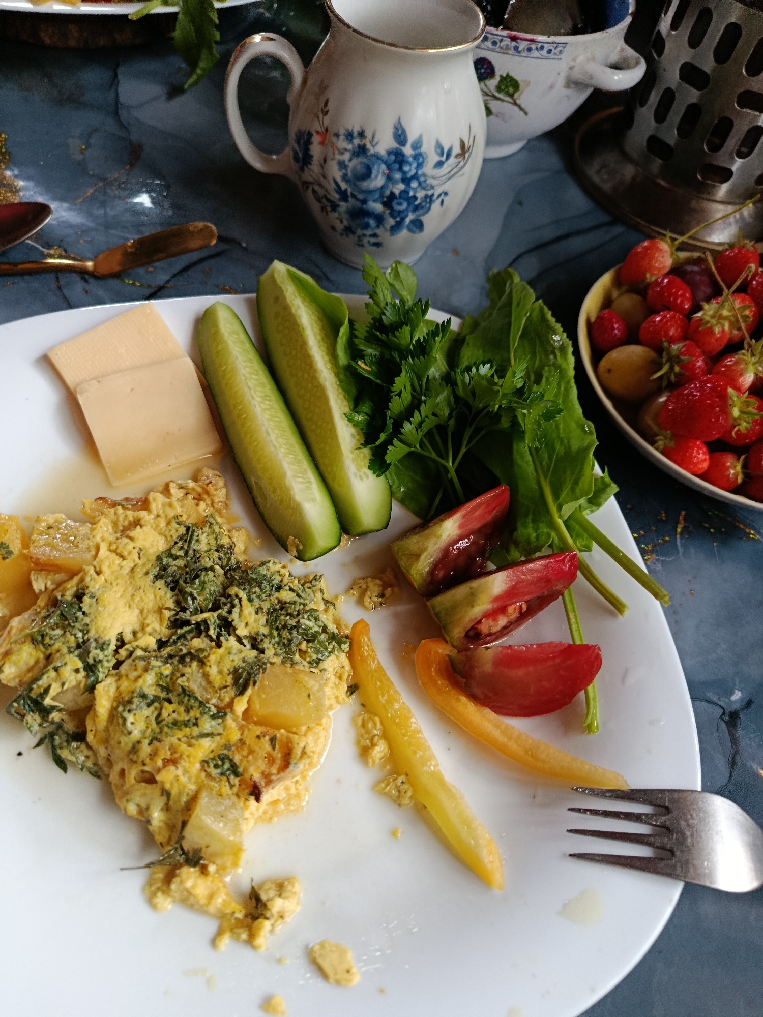 A close-up, top-down shot captures a breakfast spread on a dark, marbled surface. A white plate in the foreground holds a serving of what appears to be an omelet mixed with greens and potatoes, accompanied by two slices of cheese, two cucumber spears, and a few wedges of heirloom tomatoes and yellow bell pepper. To the right of the plate, a small bowl overflows with ripe strawberries and a few grapes. In the background, a white pitcher with blue floral detailing and a metal utensil holder with a basket weave design are visible. A single fork rests on the plate. The overall scene suggests a rustic, homemade meal. The setting is not identifiable as a specific city or location.