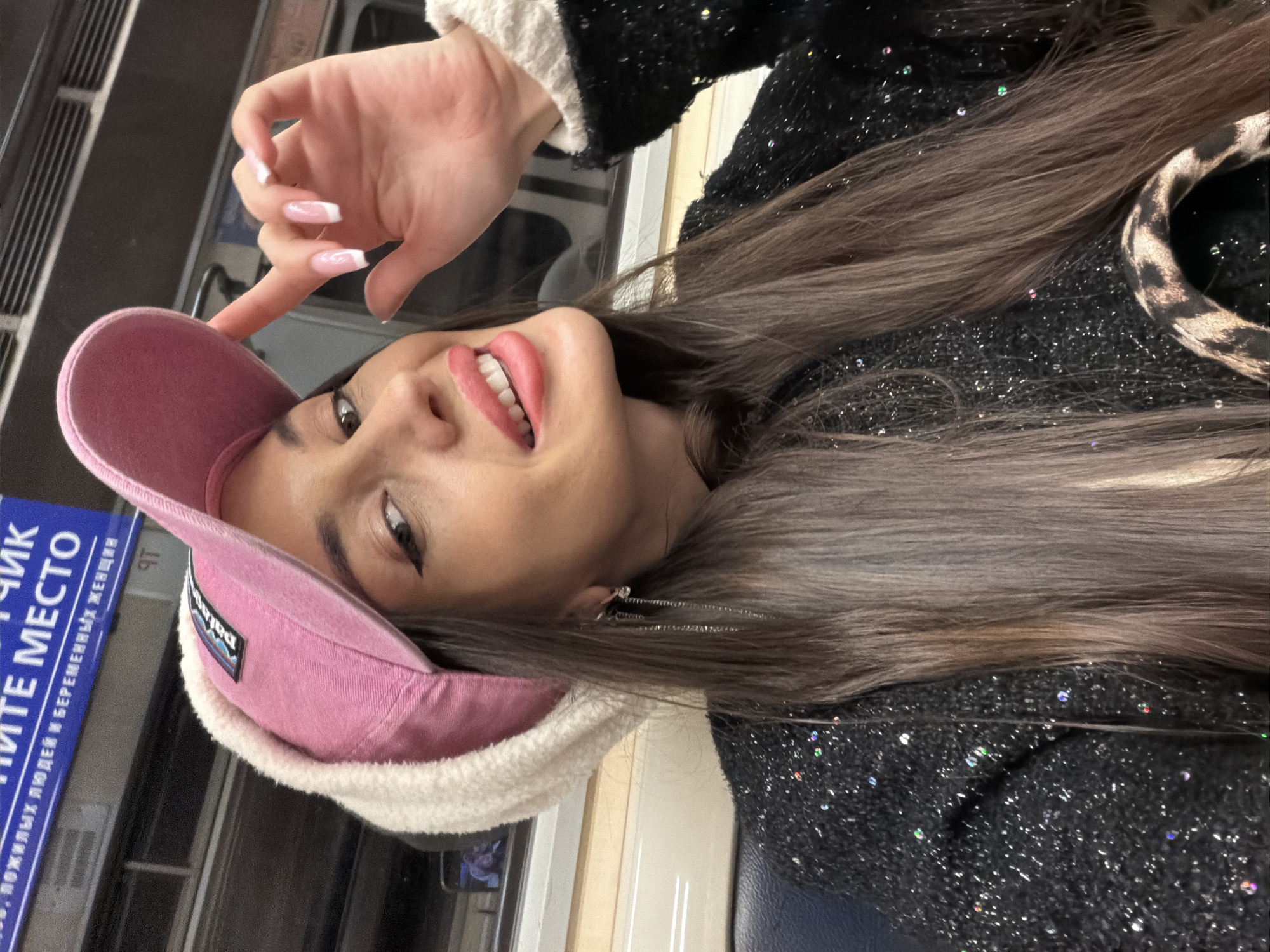 A woman is pictured inside what appears to be a public transportation vehicle. The signage is in Russian, and the image was likely taken in Saint Petersburg, Russia. She wears a pink baseball cap with a white, fluffy brim, a dark, textured jacket, and has long, light brown hair. She is making a hand gesture, possibly pointing to the hat, and is smiling. The background shows the interior of the vehicle, including what appears to be metal ventilation grates and the edge of a blue sign.